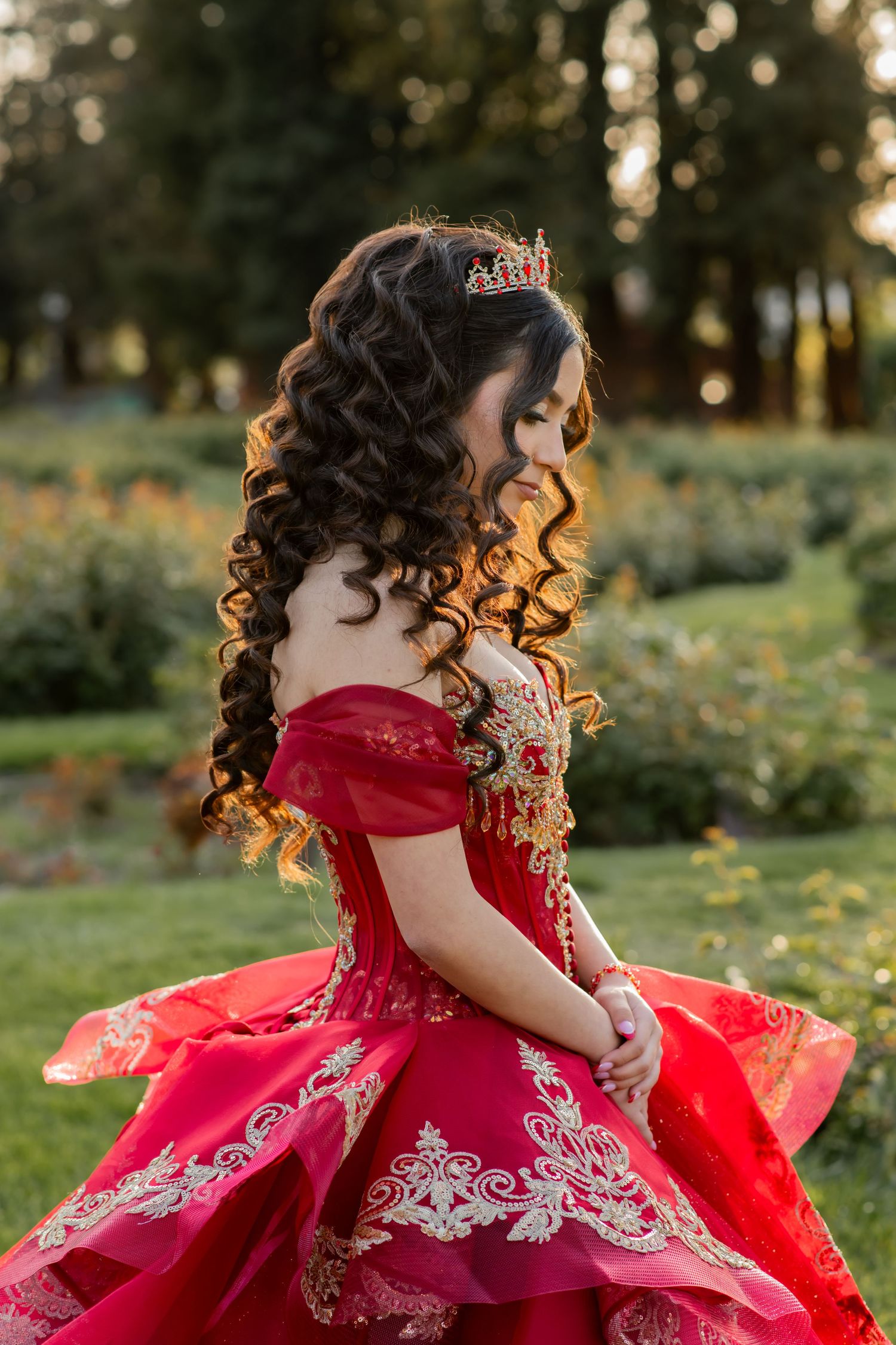 Elegant red ball gown with silver embroidery and off-shoulder design photographed in a garden during golden hour.
