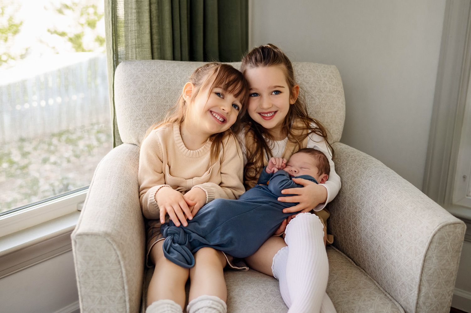 Two proud sisters sitting together holding their newborn baby sibling during a lifestyle newborn photography session