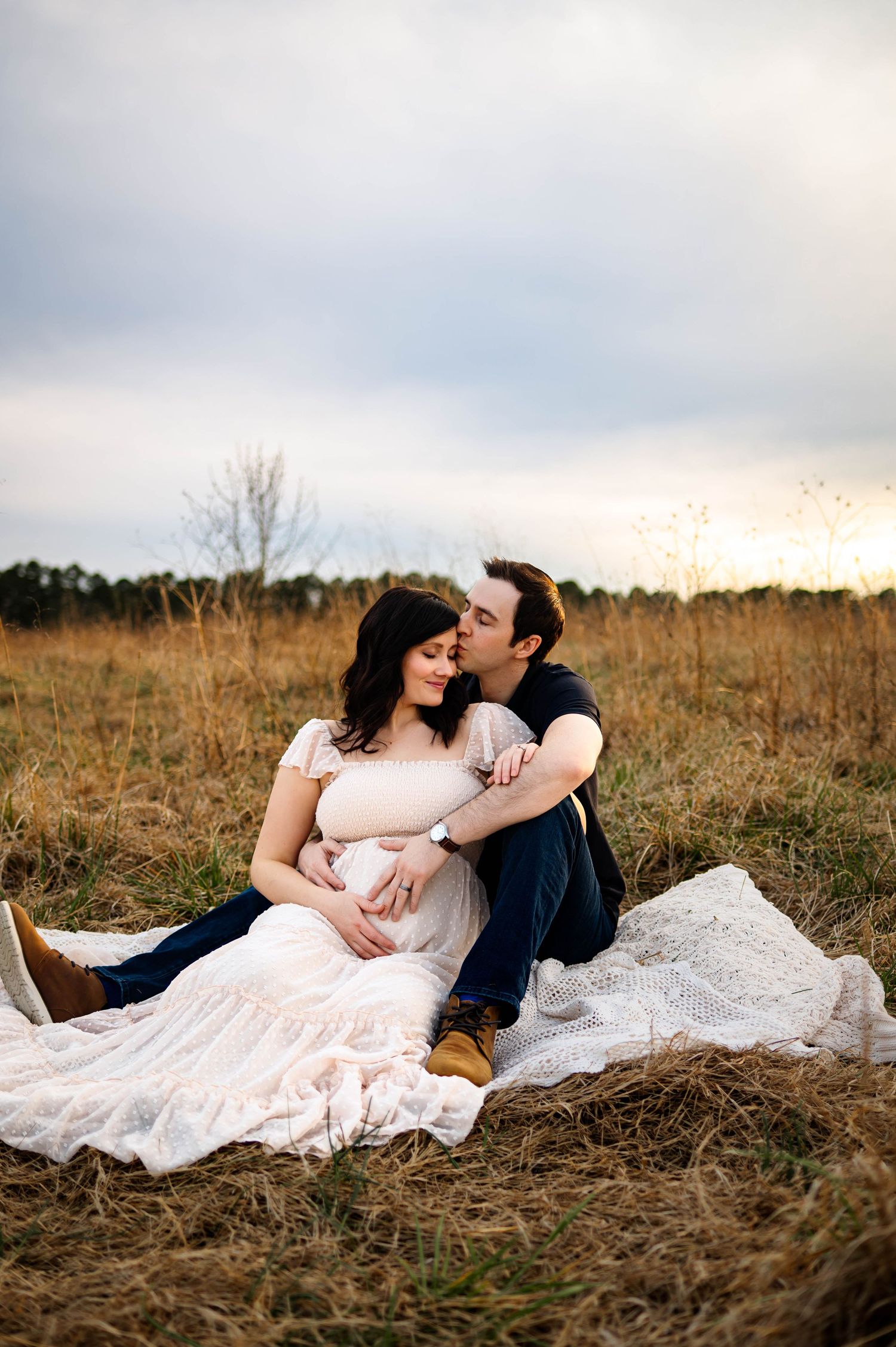 A romantic maternity photoshoot in a golden field at sunset with a couple sitting on a white blanket with Emma Branch Photography