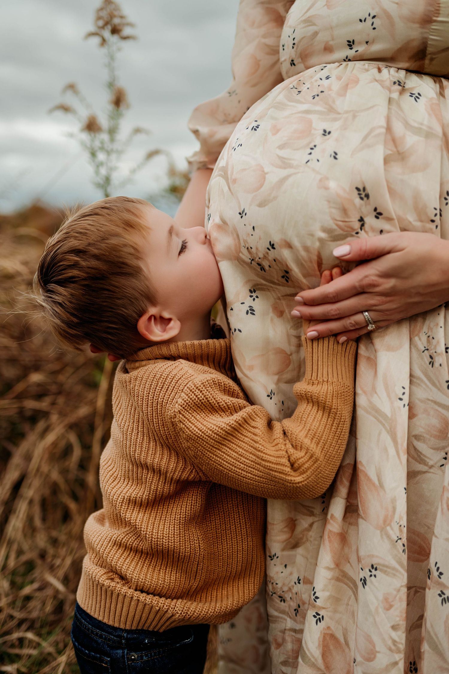 Brother in brown sweater hugging a pregnant mother wearing a floral dress and kisses belly for the best maternity photographer raleigh