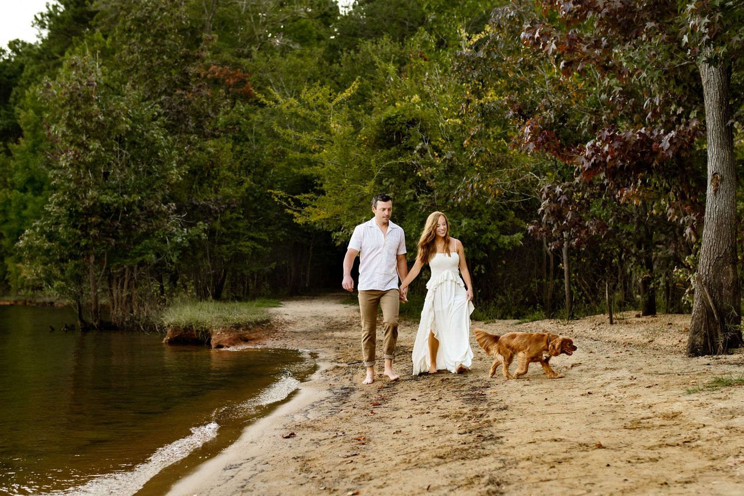 A couple in white outfits walks with their dog along a sandy beach by a lake surrounded by lush forest with a family photographer in Raleigh