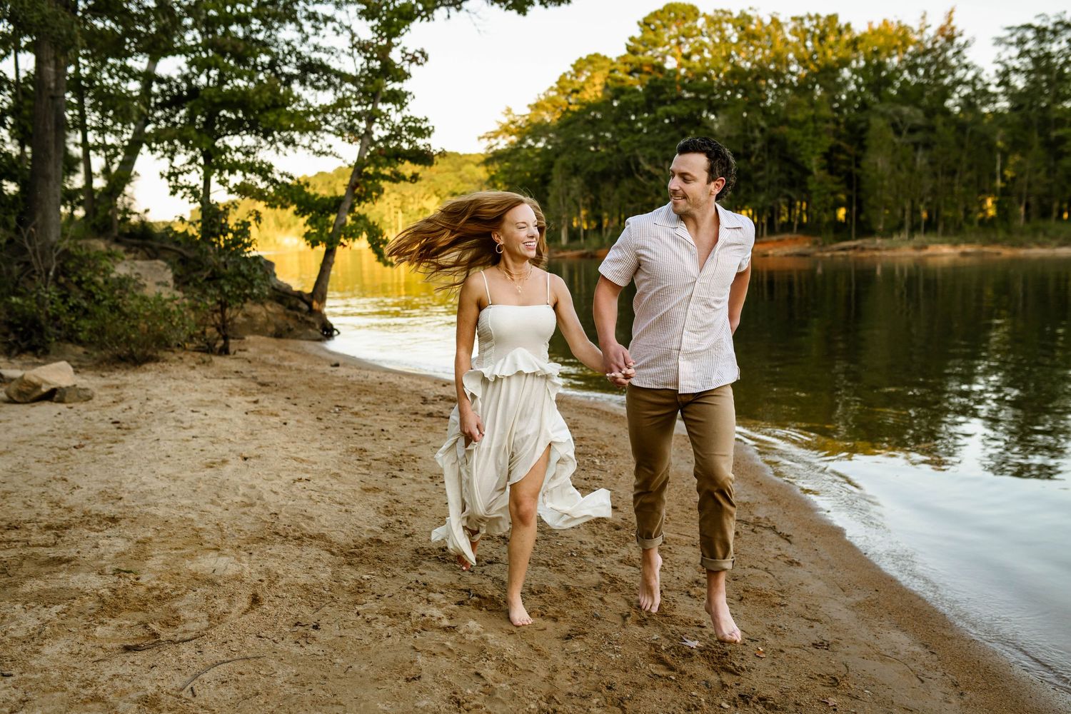 A couple walks along a scenic beach shoreline at sunset holding hands in white and casual attire with newborn photographer in Apex NC