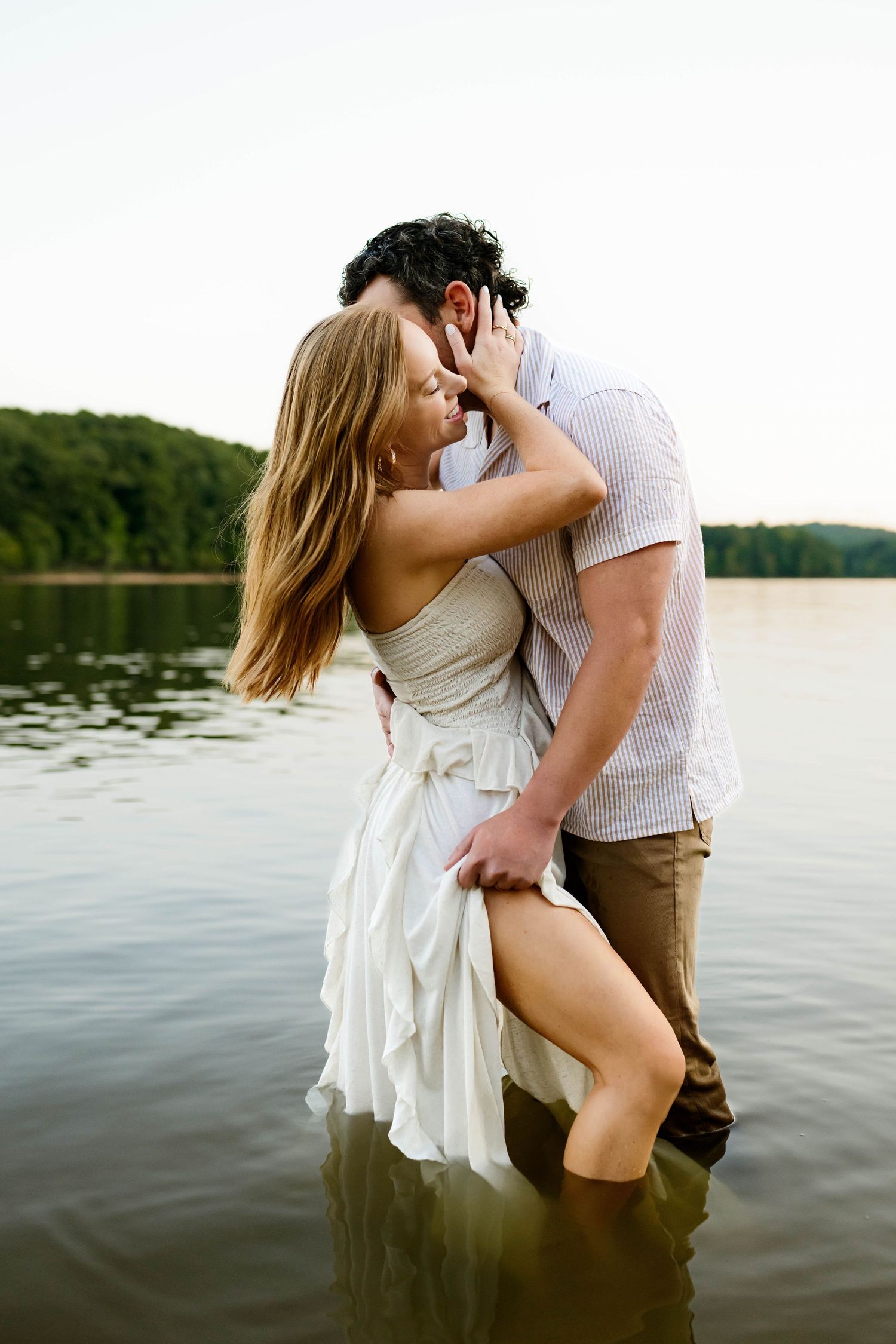 Romantic couple embraces while standing in lake water at sunset, her white dress flowing in the gentle breeze with Emma Branch Photography