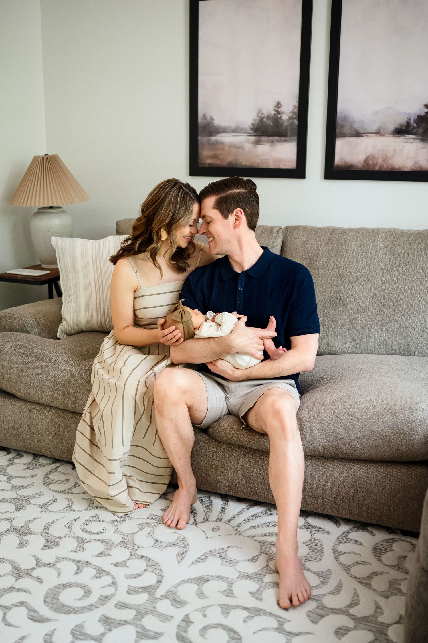 New parents sit together on couch in neutral living room while holding newborn baby during a family photography session in Raleigh NC