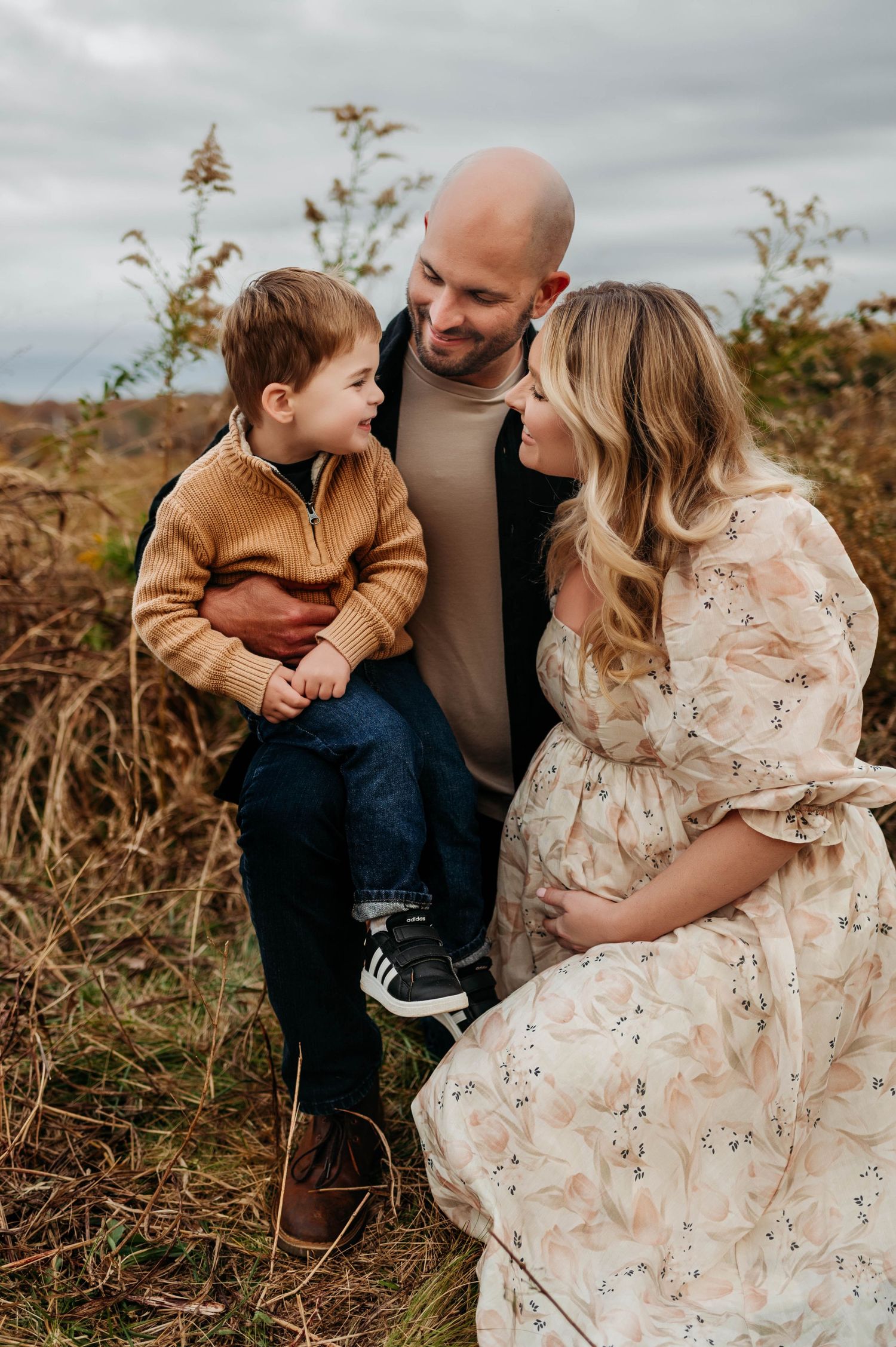 A family shares a tender moment outdoors on an autumn day, with a floral dress and brown sweater with maternity photographer chapel hill