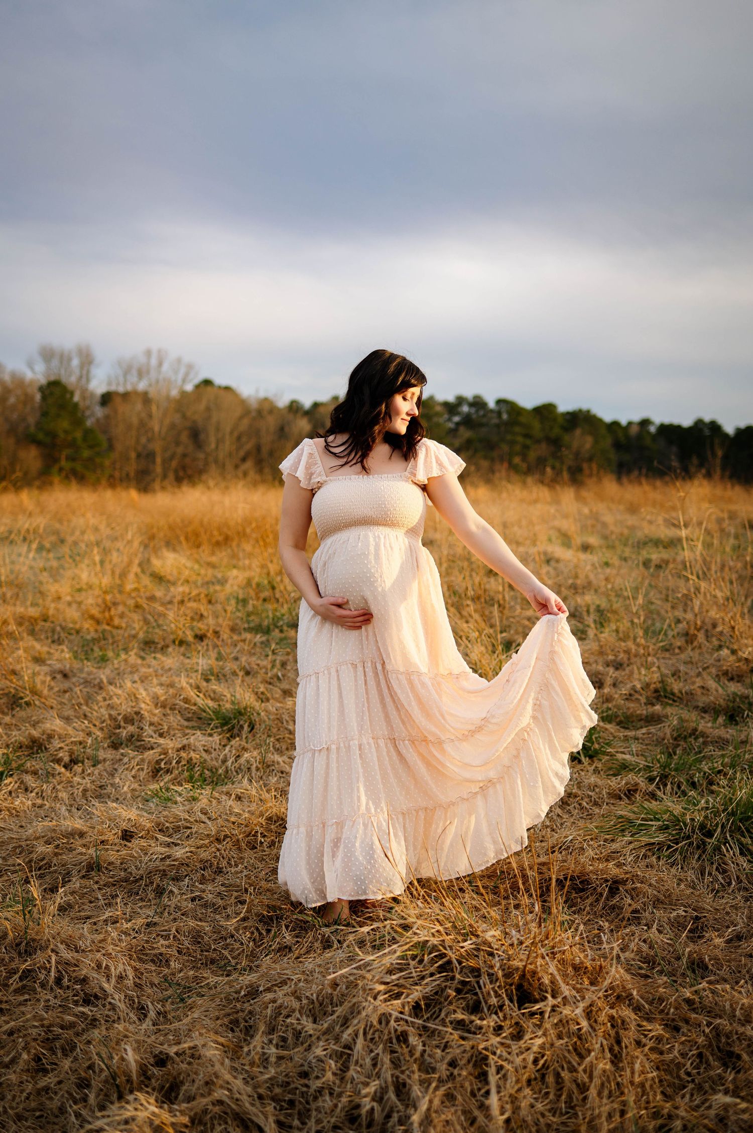 A flowing cream-colored maxi dress catches the golden light in a rustic field during a maternity photoshoot in Wake Forest, NC