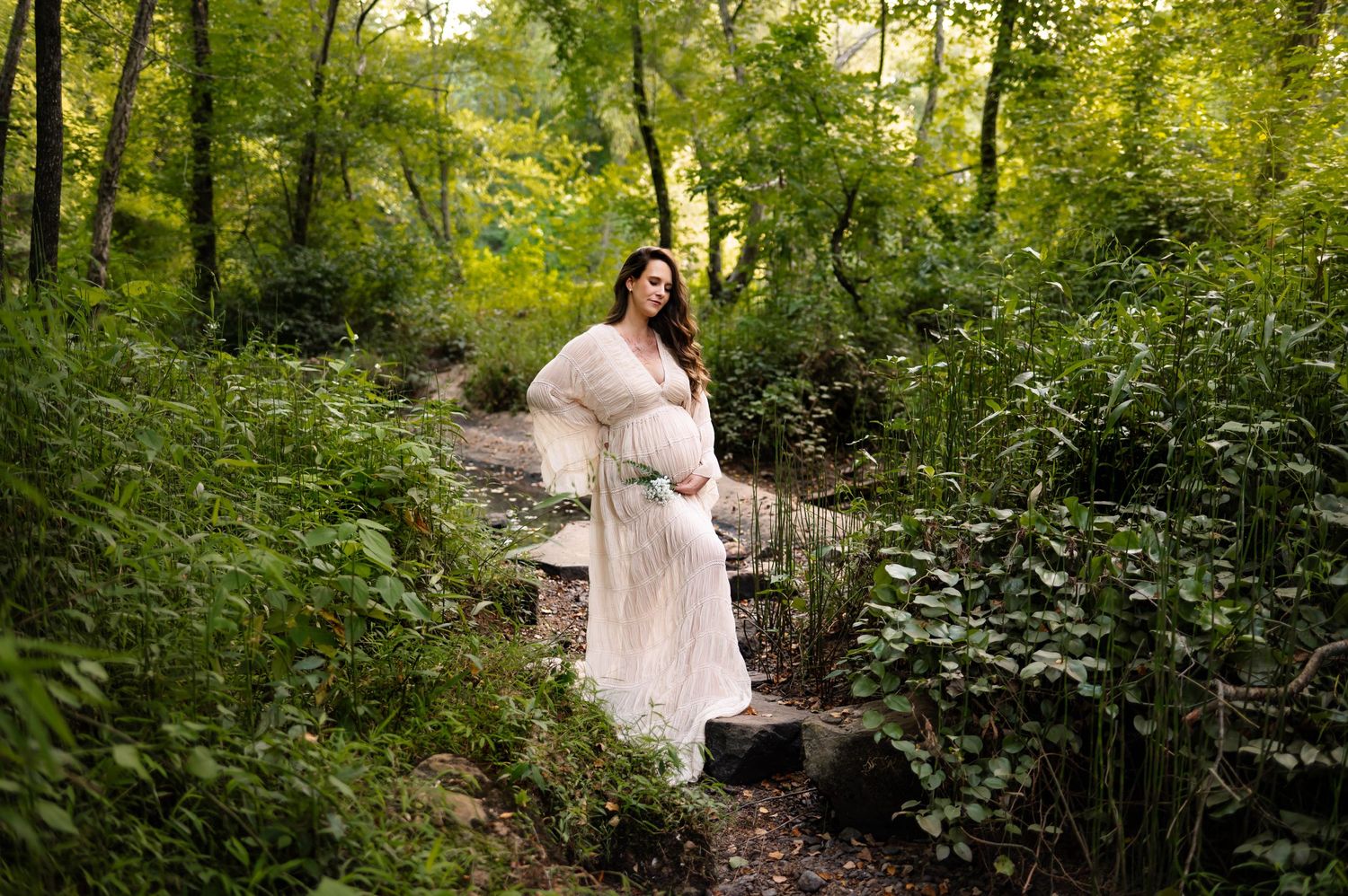 A mother in a flowing white dress stands on stones surrounded by lush green forest foliage with raleigh nc maternity photographer