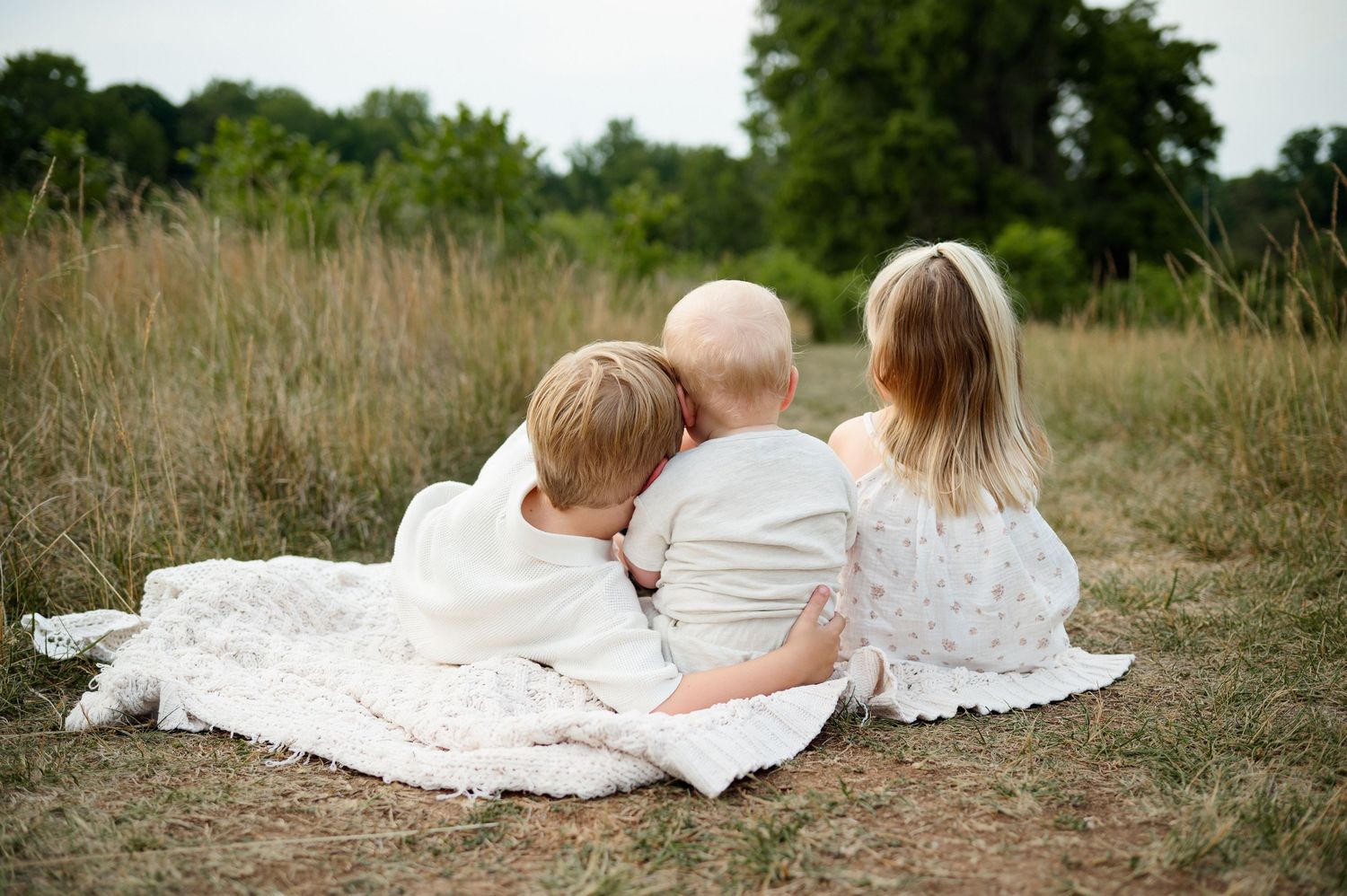 Three siblings sitting together on a blanket in a grassy field during a family photography session in Raleigh NC