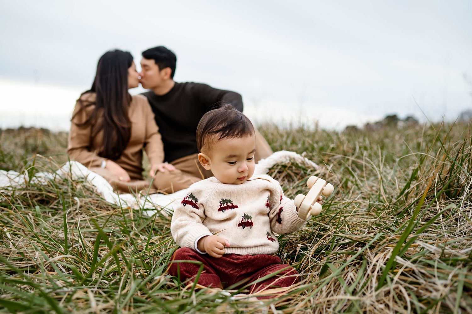 Toddler sitting in a grassy field playing while parents sit together in the background during a Raleigh family photography session