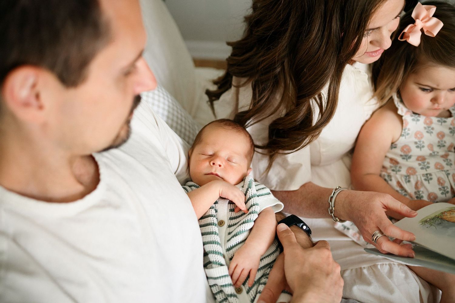 Parents reading with their toddler daughter while their newborn baby sleeps in dad's arms during an in-home newborn photography session