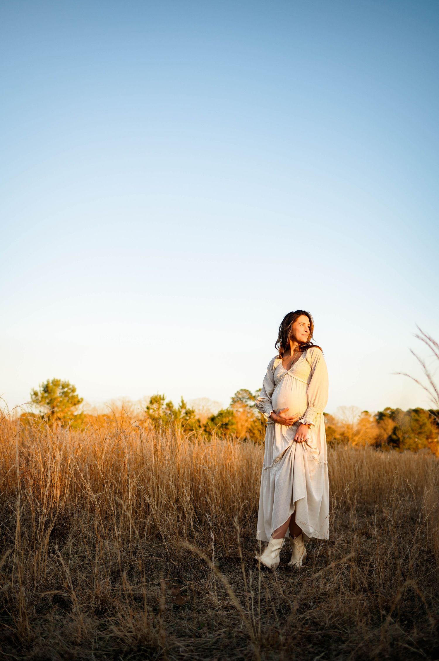 Mother in a flowing white dress stands in a golden field at sunset while cradling their pregnant belly with apex maternity photographer