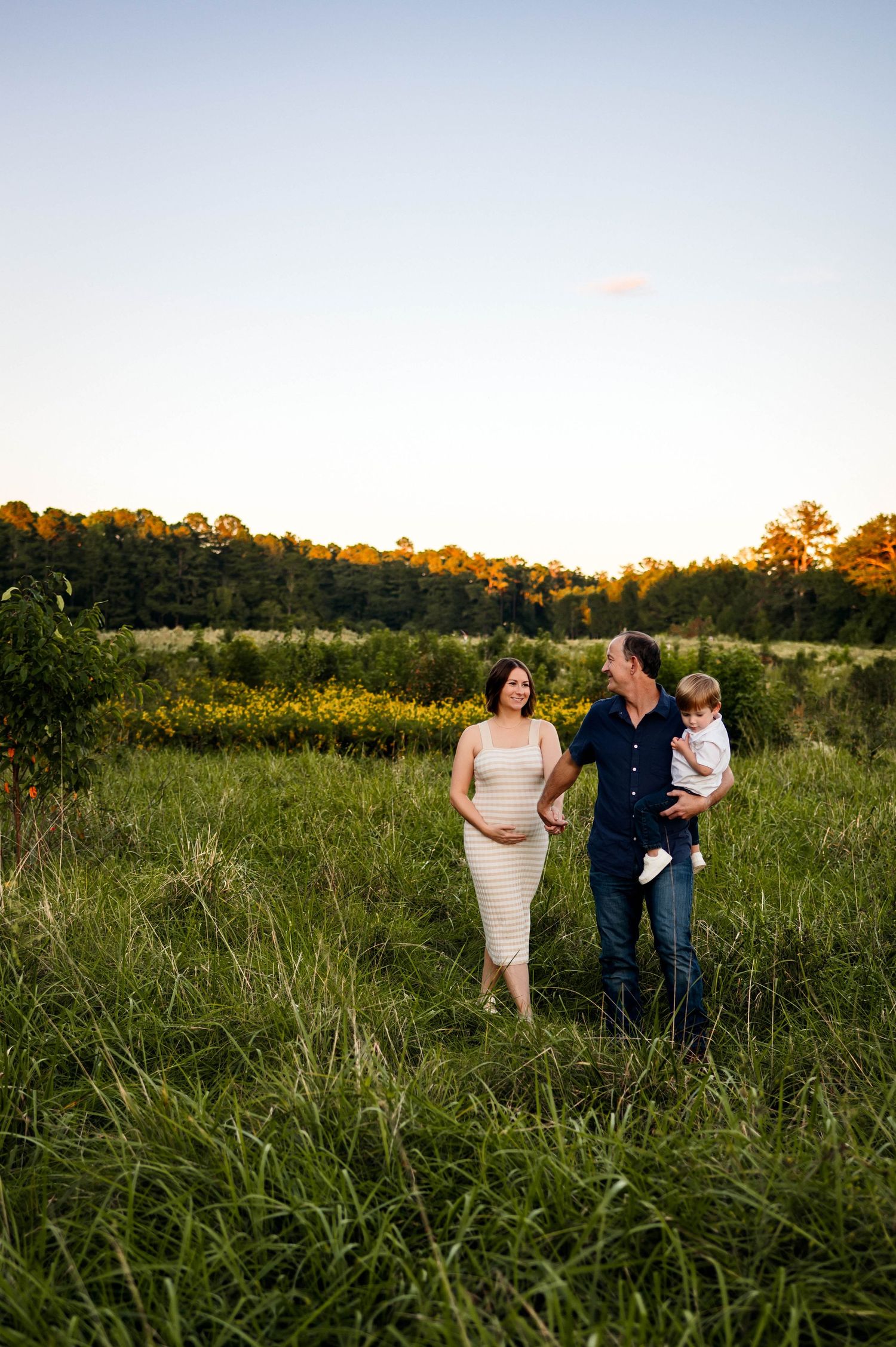 A family walks together through a golden grassy field at sunset with trees lining the horizon with Raleigh NC maternity photographer