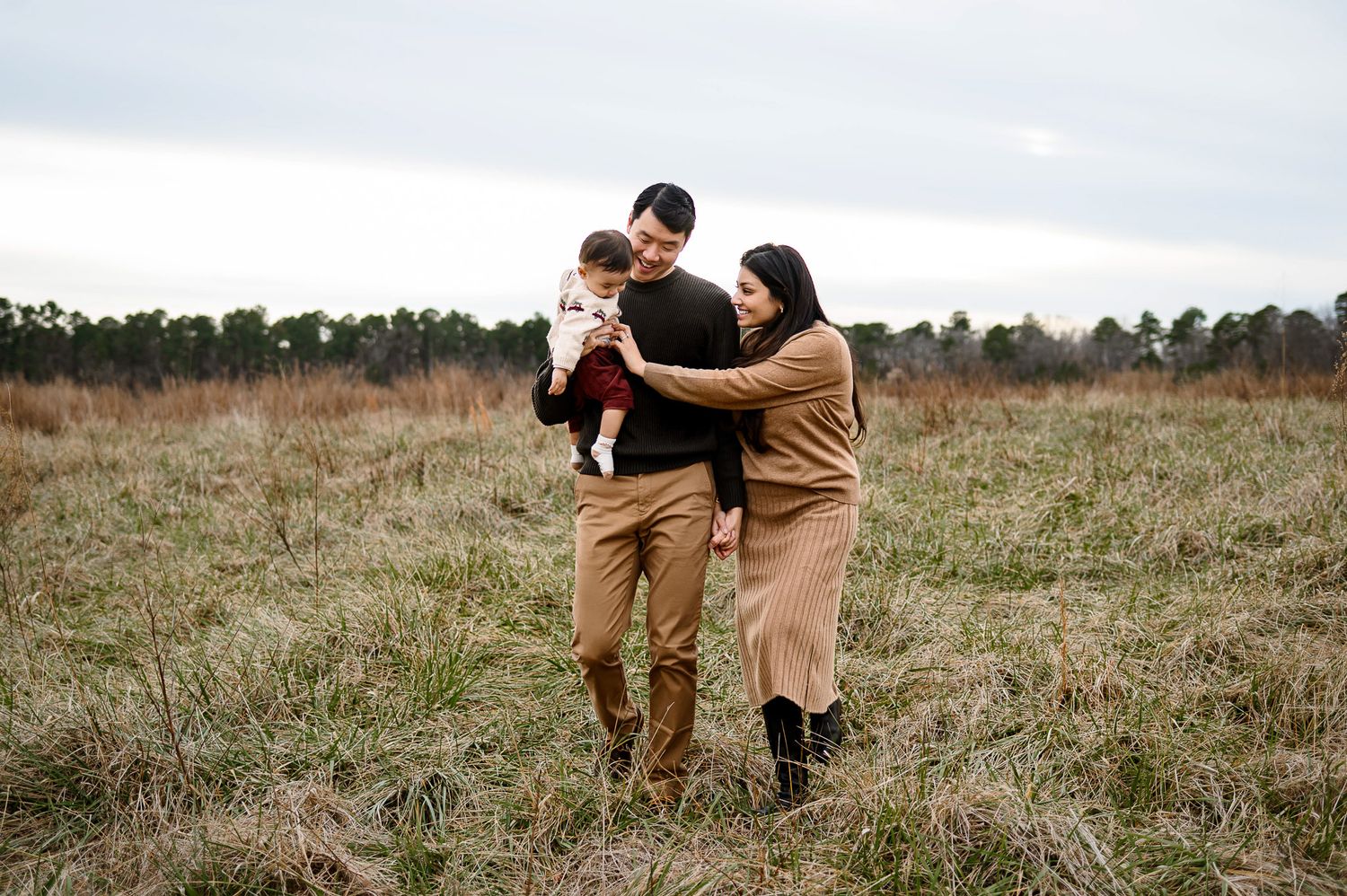 Parents walking together with their baby through a grassy field during a family photography session in Wake Forest NC