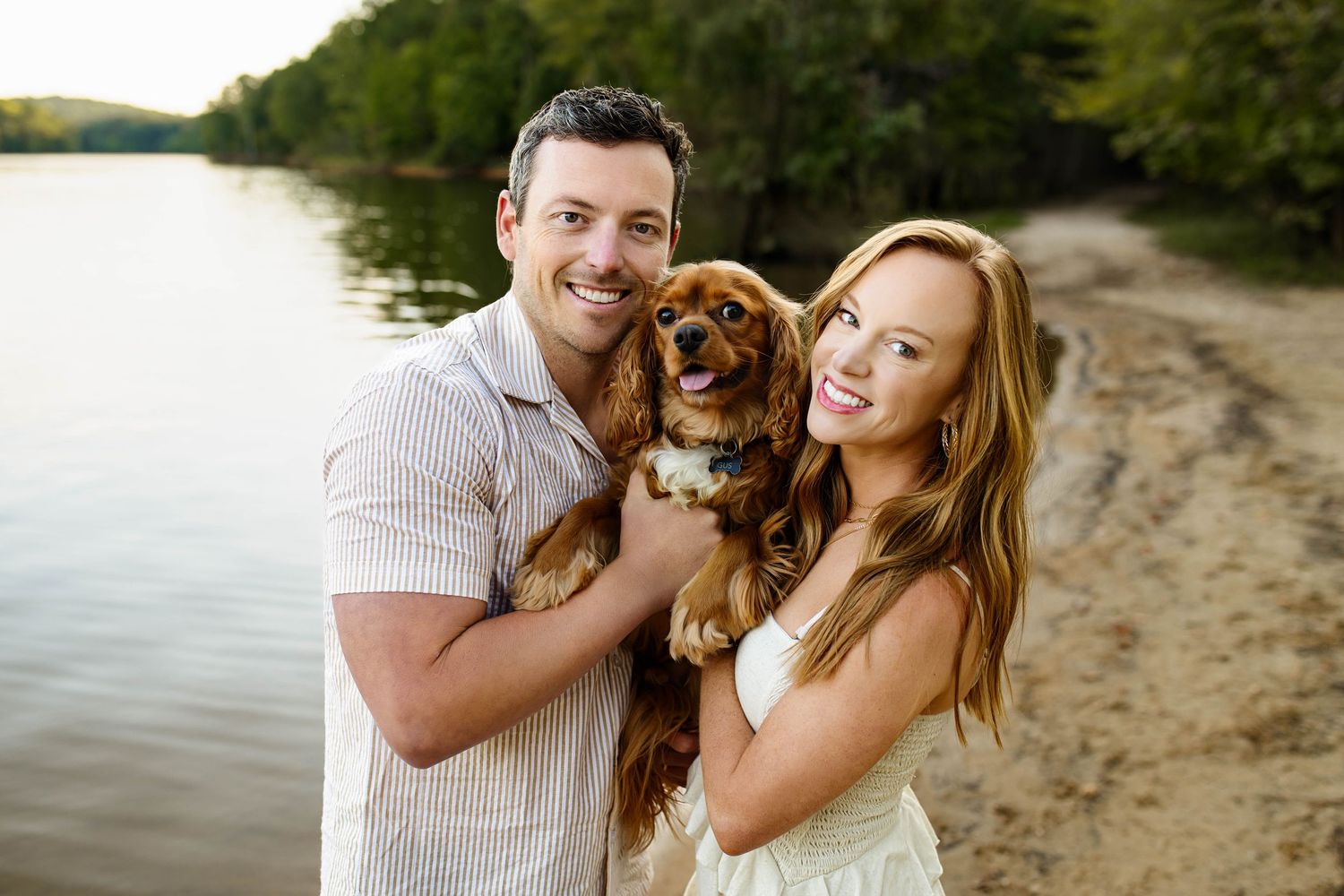A couple enjoys a playful moment with their puppy on a sunny beach beside a lake in Raleigh NC with a family photographer.