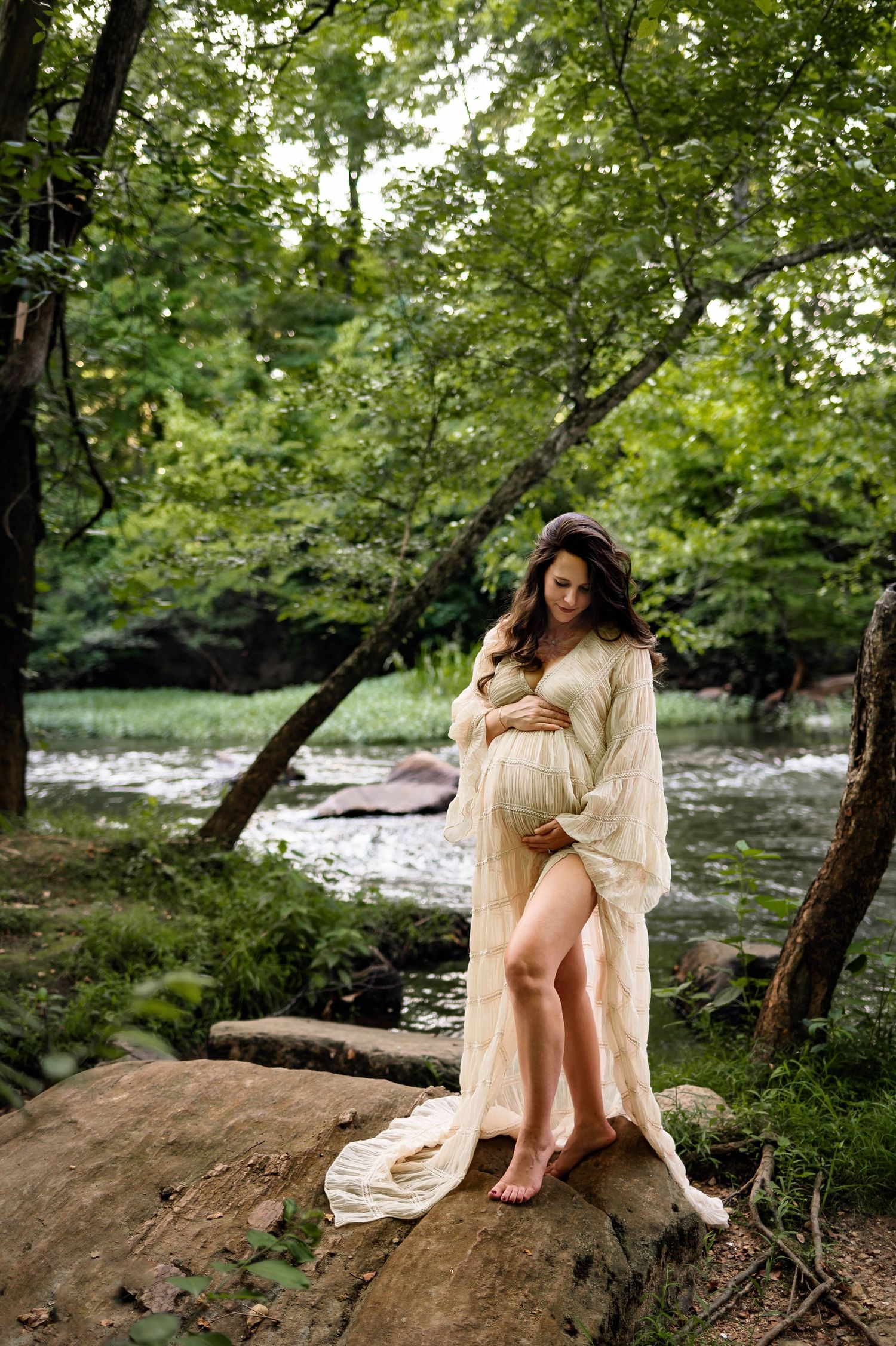 A maternity photoshoot in Wake Forest, NC by a peaceful stream and rocks and mother cradles her growing belly with maternity photographer