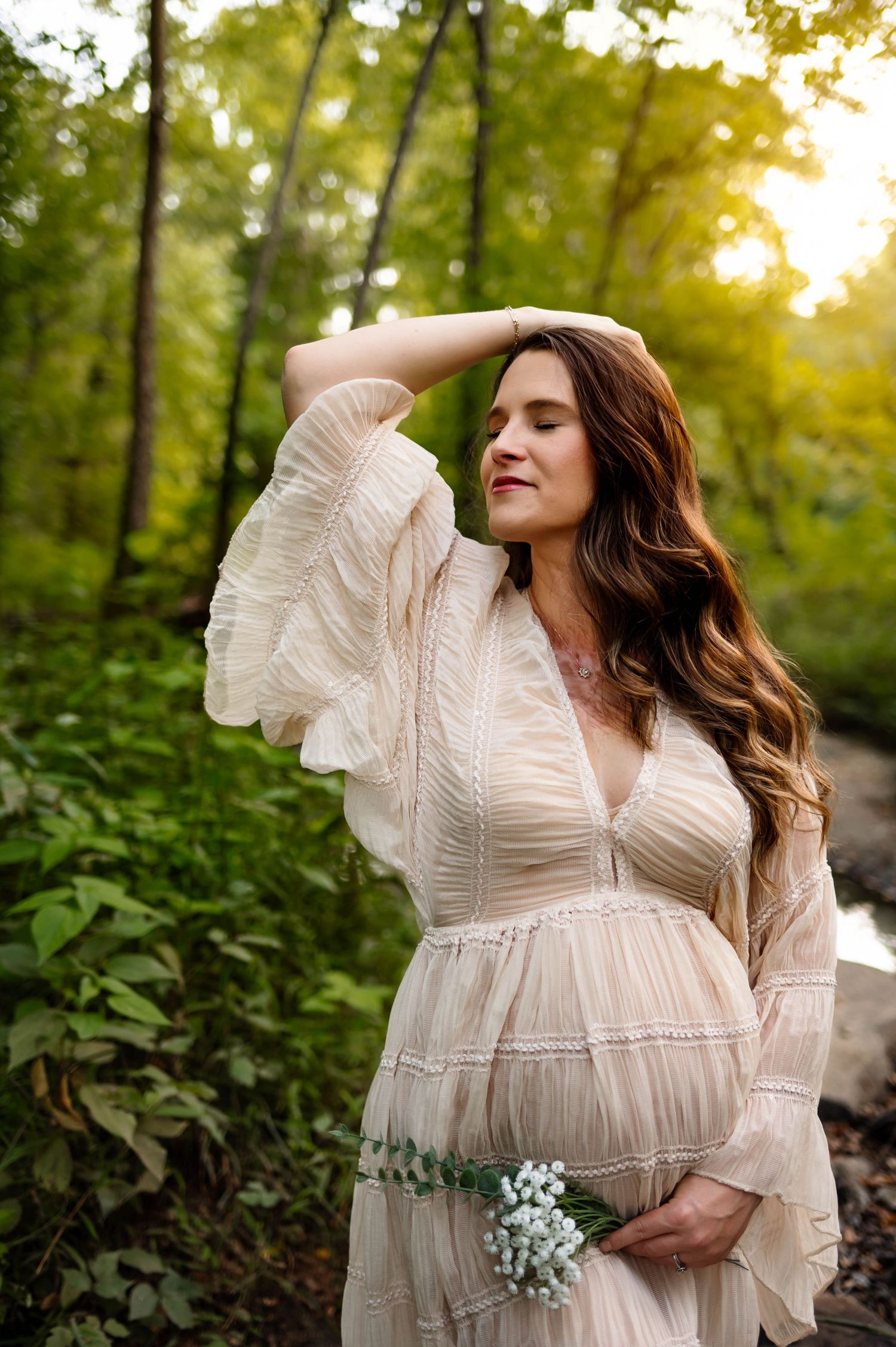 Maternity photo in a dreamy lace dress holding baby's breath flowers in a sunlit forest setting with raleigh maternity photographer