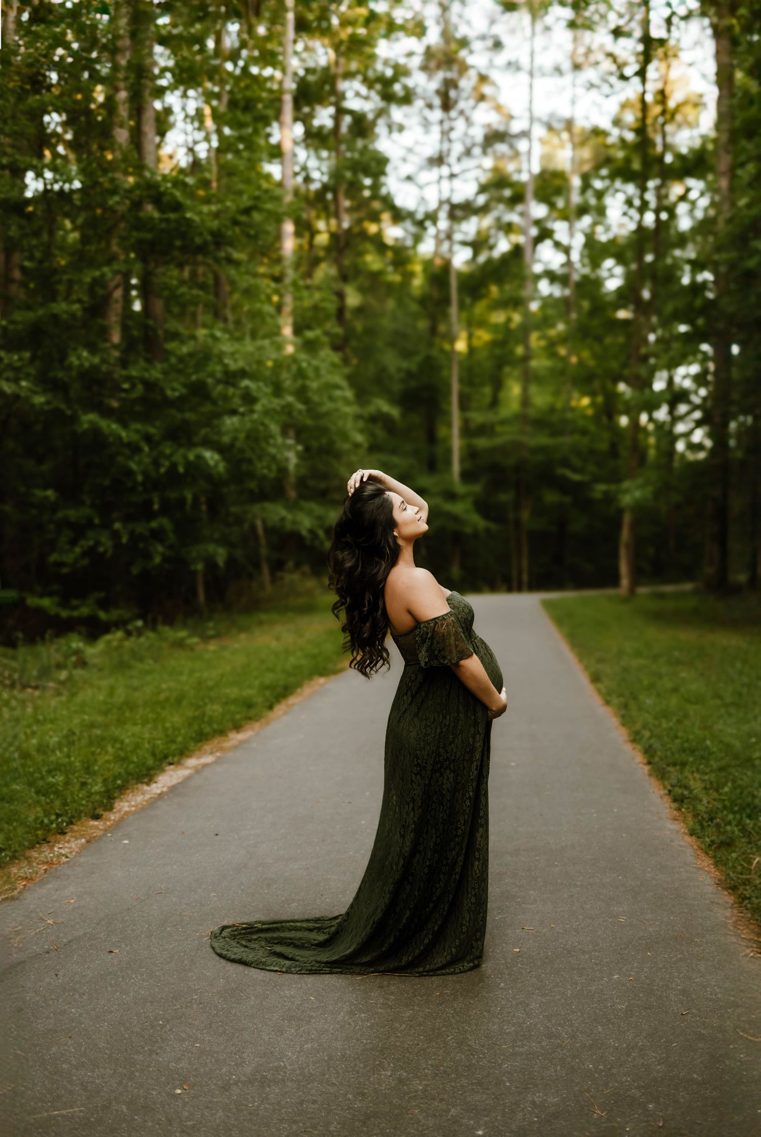Mother in flowing dark formal dress poses elegantly on a tree-lined path at golden hour for maternity photography in Wake Forest