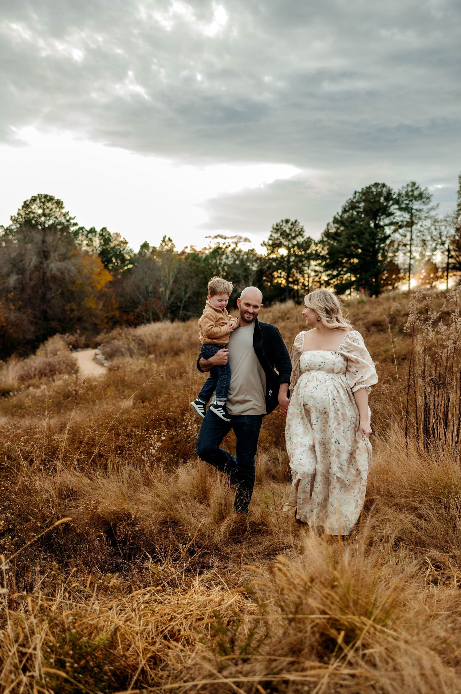 A family walks together through golden autumn grass in a field at sunset with moody clouds overhead during maternity photoshoot