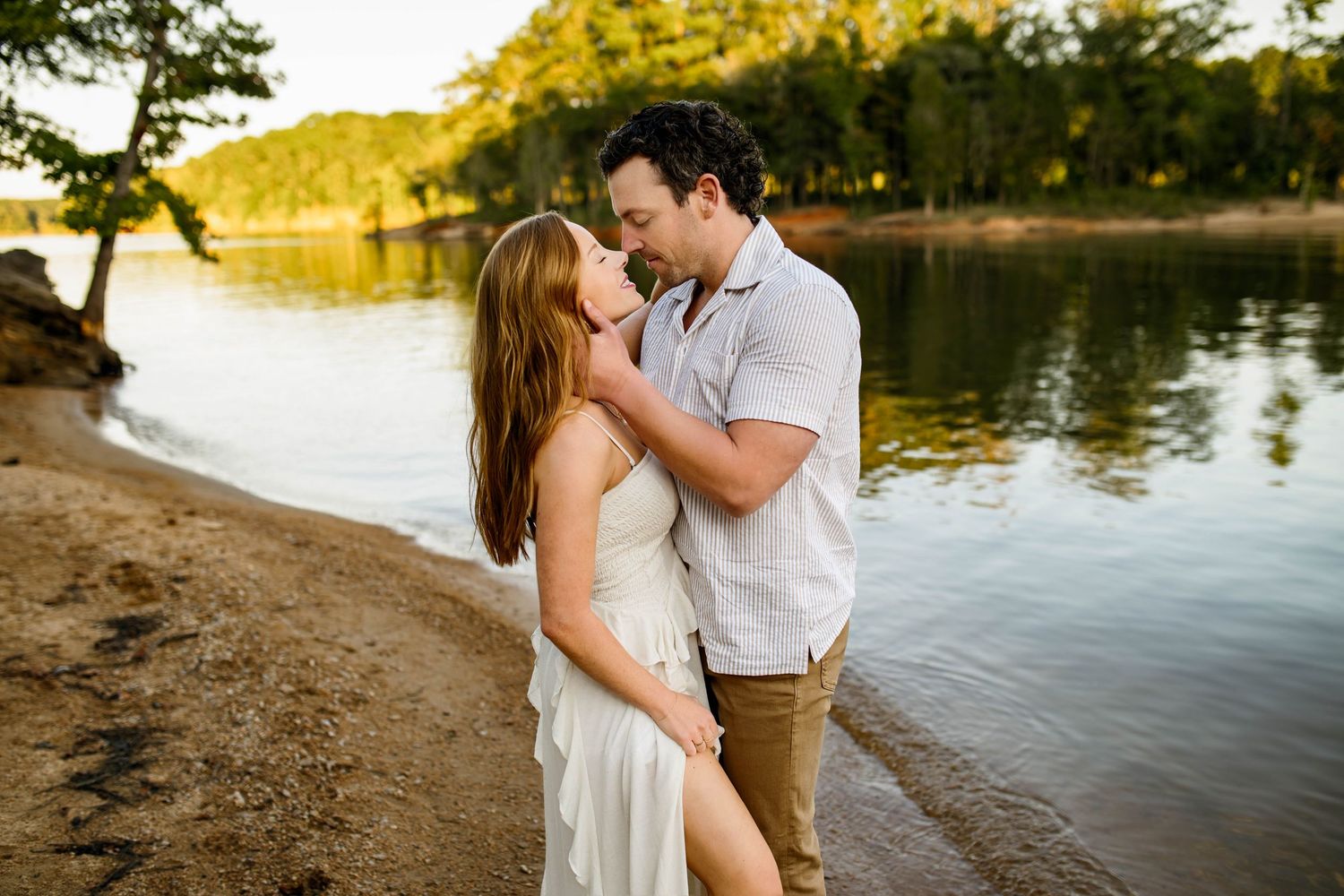A romantic couple embraces on a sandy beach at sunset beside a tranquil lake surrounded by trees with Wake Forest NC newborn photographer