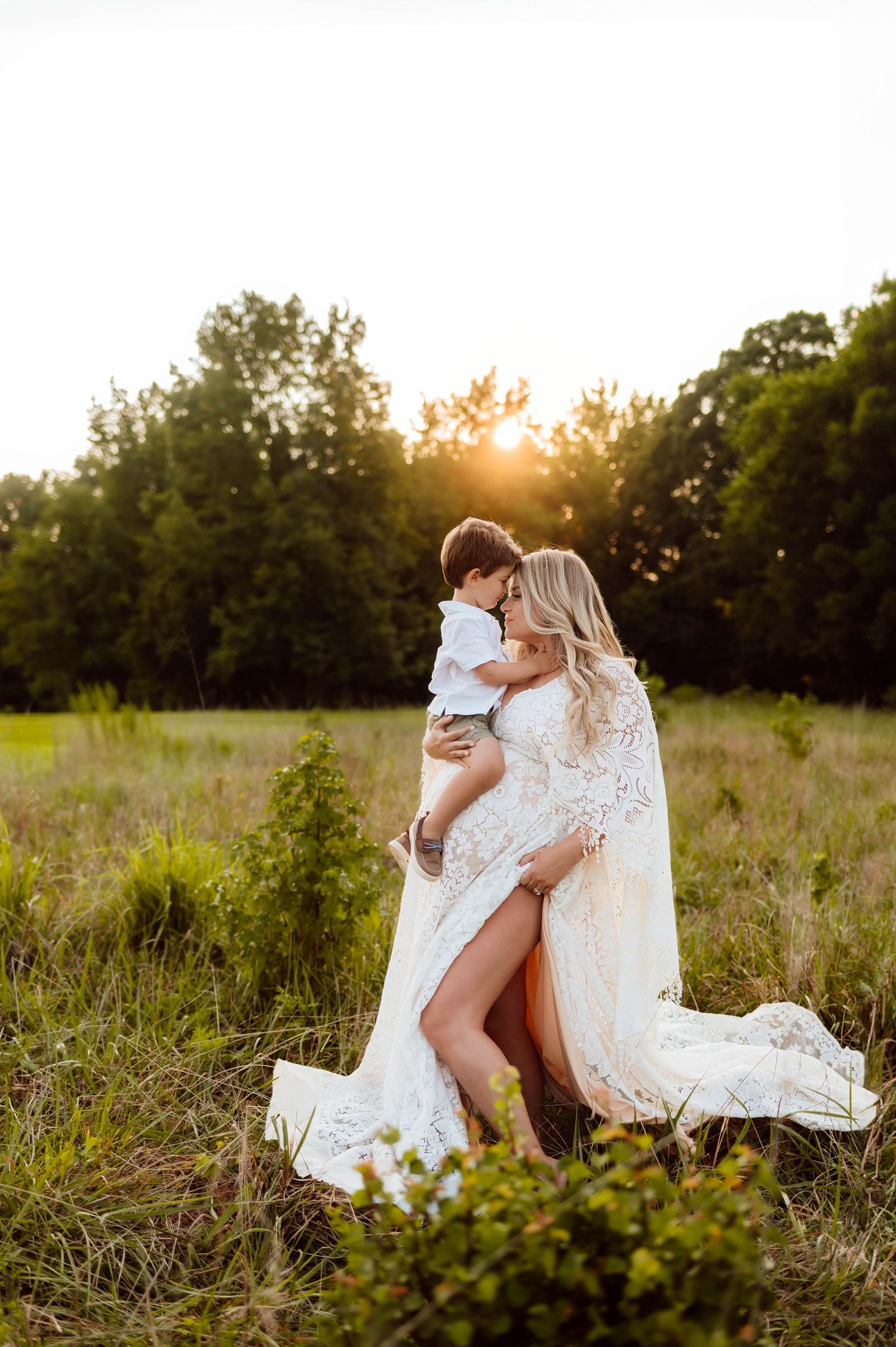 A tender moment captured in a sun-drenched field at golden hour as a white lace dress flows with maternity photographer apex