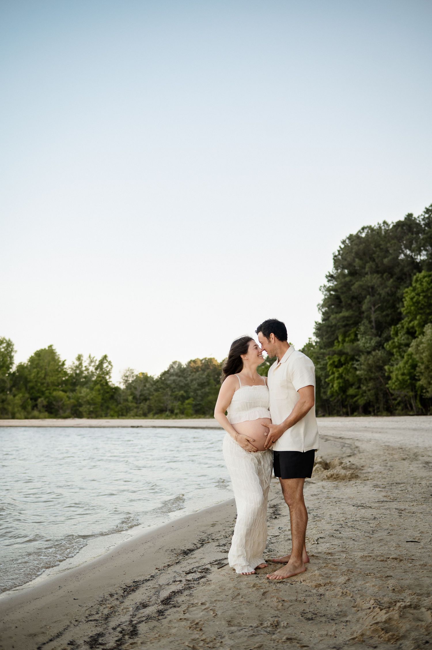Cary family photographer captures expecting parents smiling together while gently holding mother's pregnant belly along the water's edge