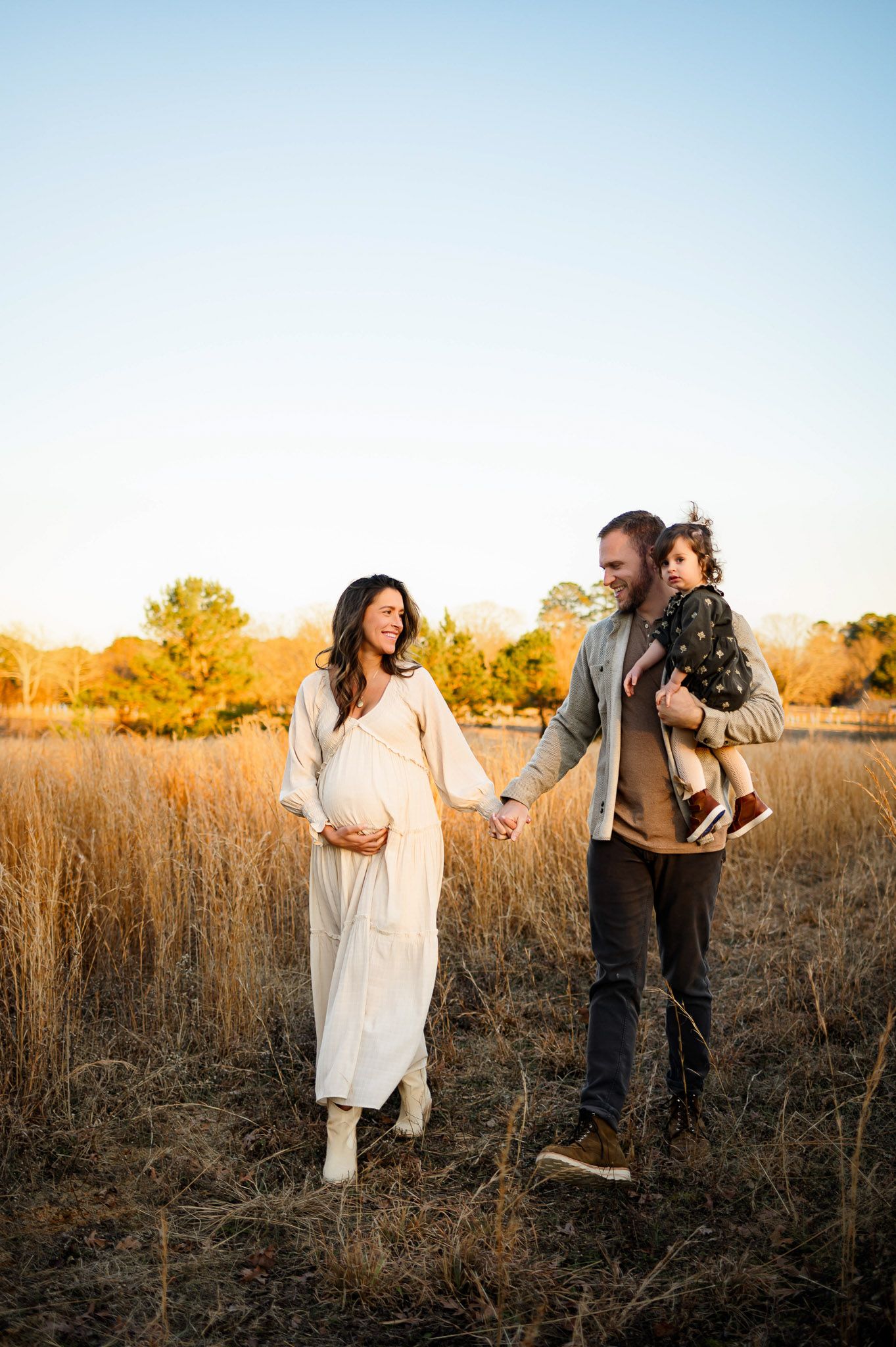 Pregnant mother walking through a golden field with her husband holding their toddler during an outdoor maternity photo session in Raleigh