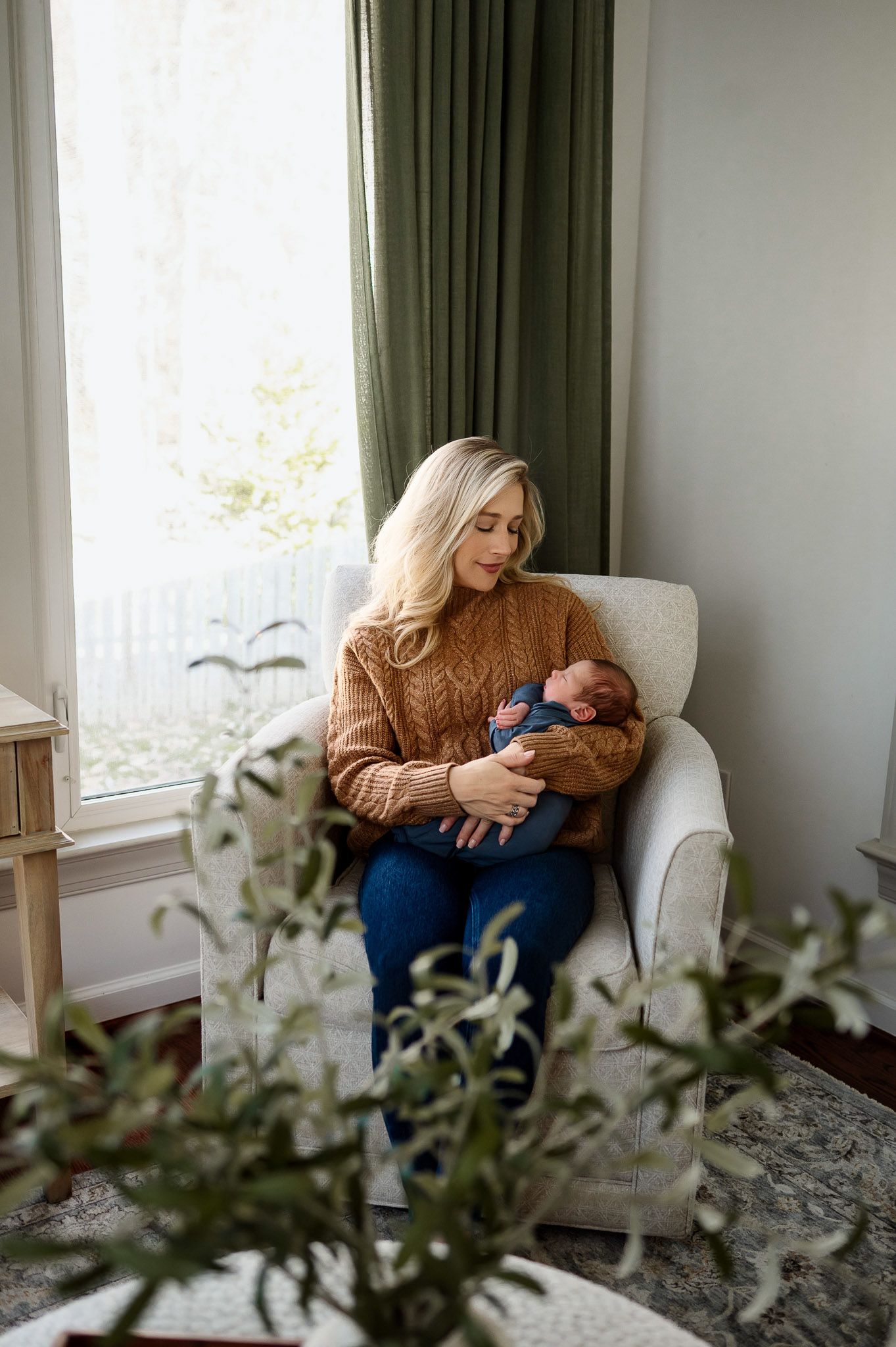 Mother sitting in a chair holding her newborn baby beside a window during an in-home Raleigh newborn photography session