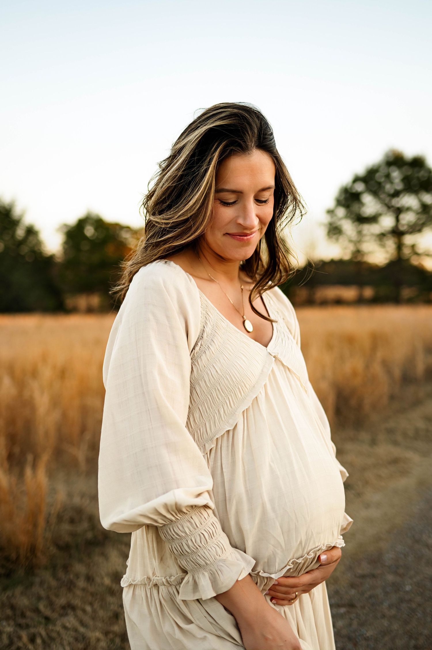 Expectant mother in white flowing dress stands peacefully in golden field at sunset cradling her belly with apex maternity photographer