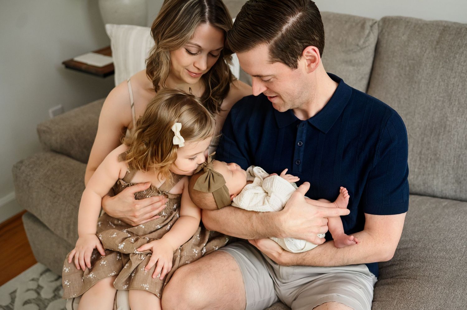 Family of four sit together on couch in their home while older sibling kisses baby during a newborn photography session in Raleigh NC