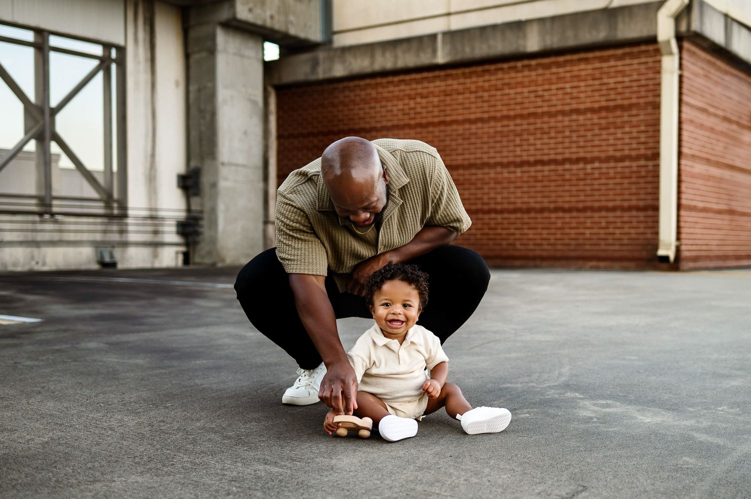 Father crouching to play with smiling son on packing deck rooftop during candid lifestyle family photography session in downtown Raleigh NC
