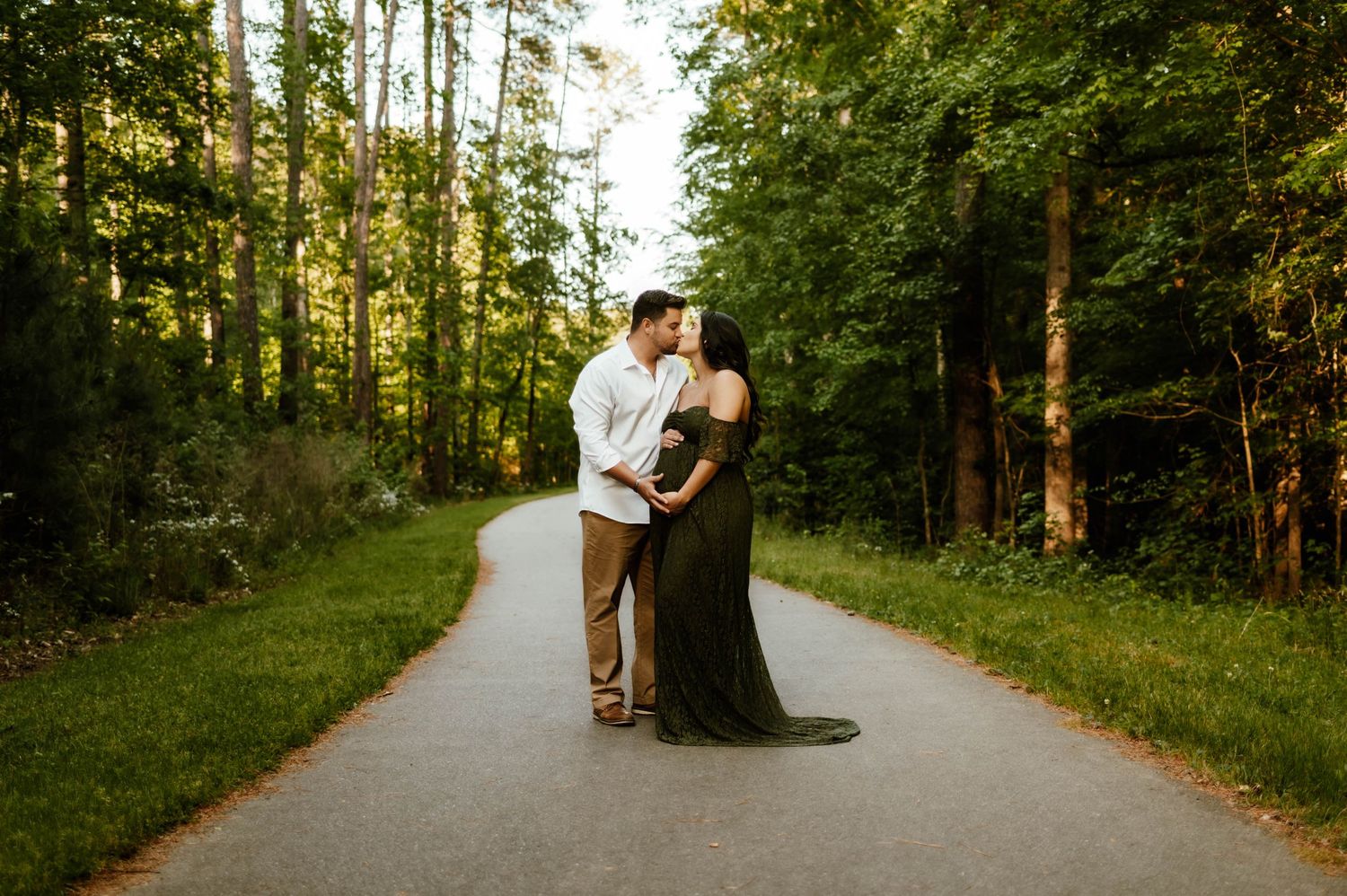 A romantic couple embrace on a winding path during golden hour sunset surrounded by tall green trees for maternity photographer durham