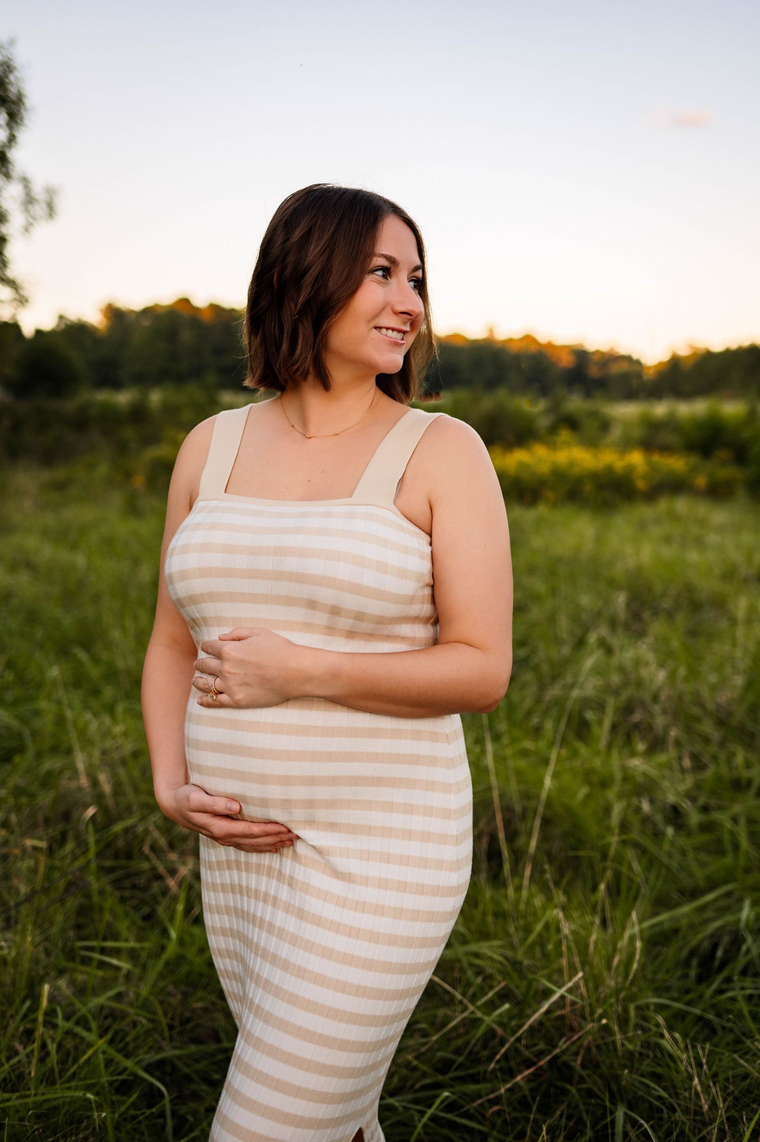 A mother in a striped beige dress stands in a golden field at sunset while smiling with maternity photographer raleigh