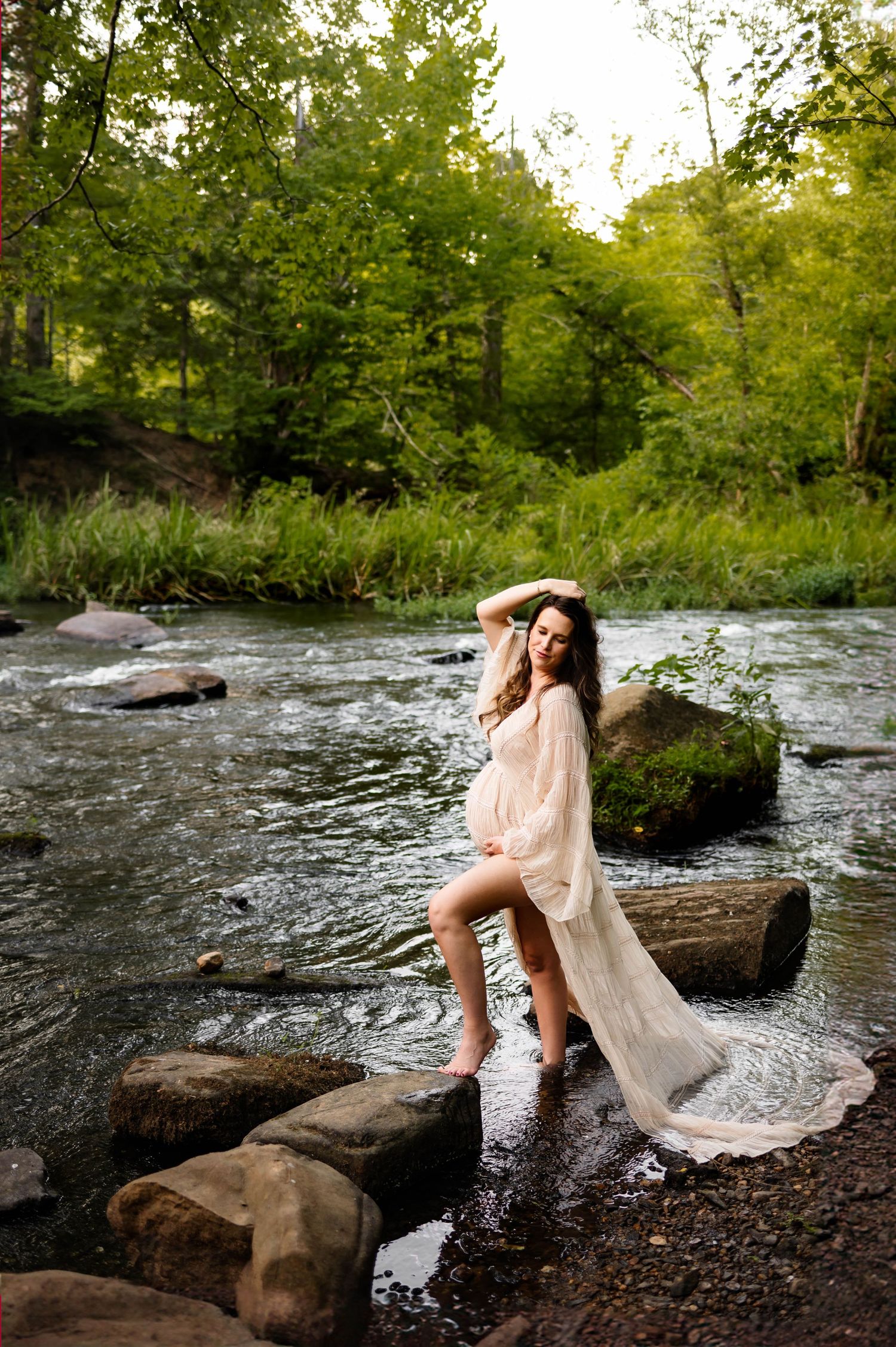 Expecting mother stands in river waters in a lush forest setting while tossing her hair during session with raleigh maternity photographer