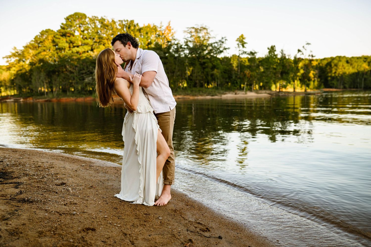 Couple embraces on a beach at sunset with golden light reflecting off a serene lake in the background with newborn photographer in Raleigh