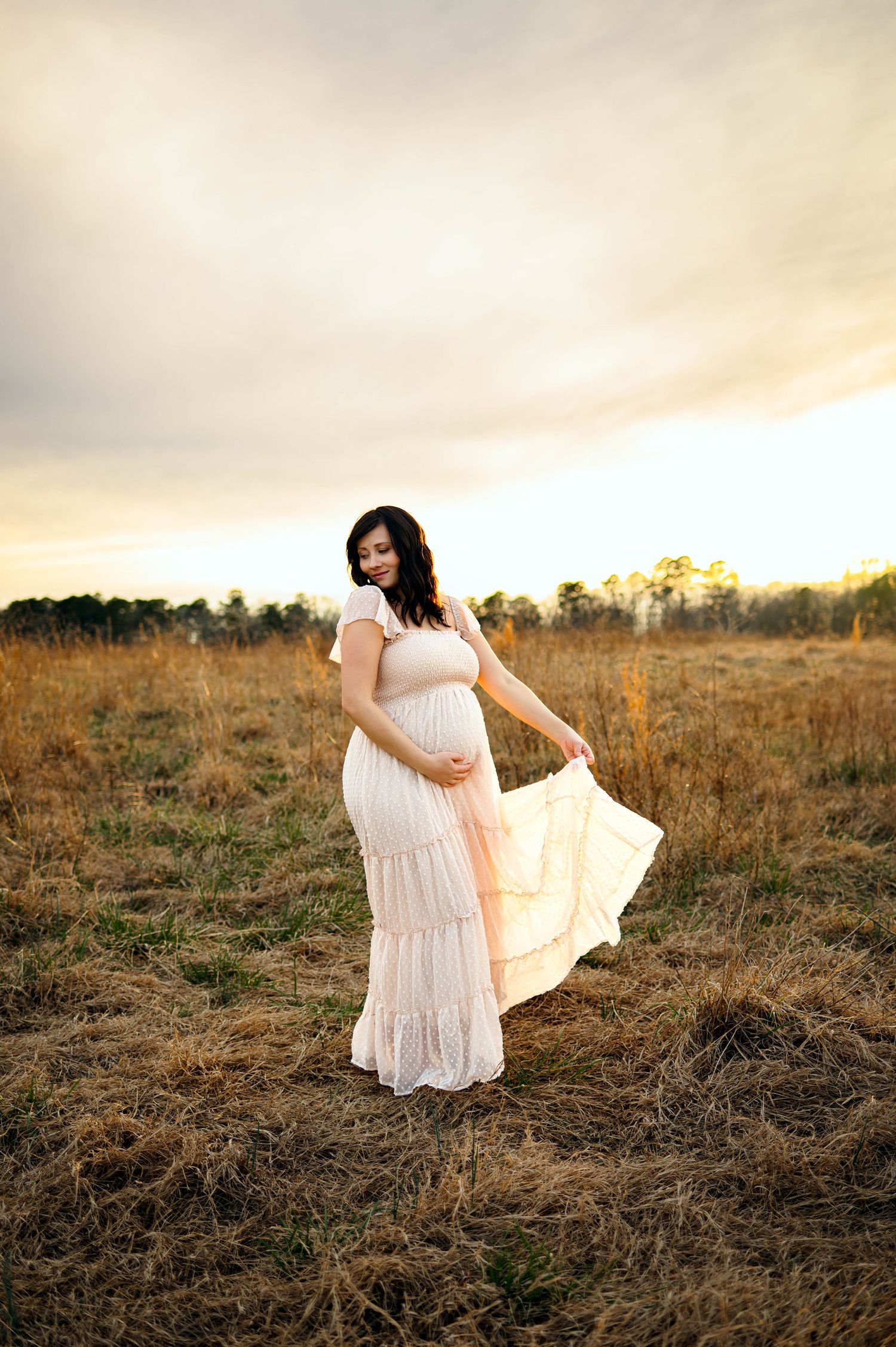 A pregnant woman in a flowing white dress stands in a golden field at sunset with raleigh maternity photographer