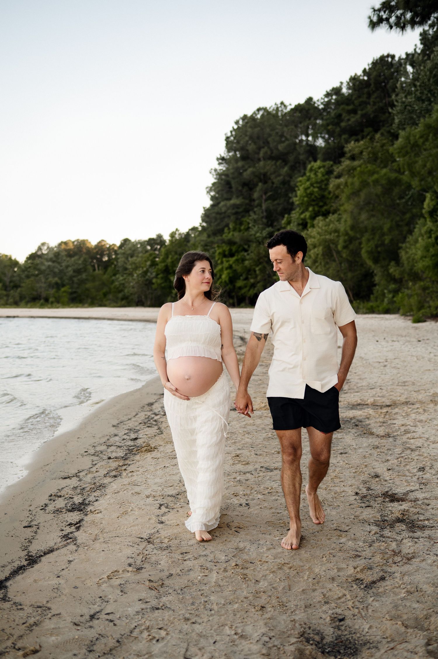 Expecting parents walk along the water's edge of Jordan Lake in Raleigh during a maternity photoshoot with Emma Branch Photography