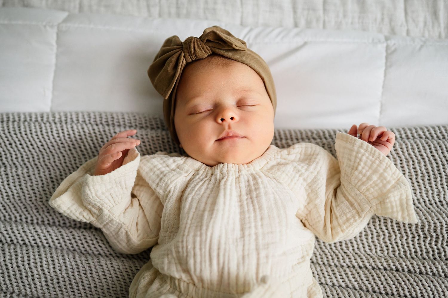Newborn baby wearing a cream top and brown bow sleeps peacefully on bed during a newborn photography session in Raleigh NC