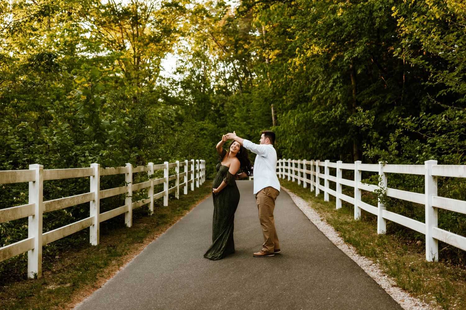 A couple shares a romantic moment on a pathway lined with white fences and lush green trees with durham maternity photographer