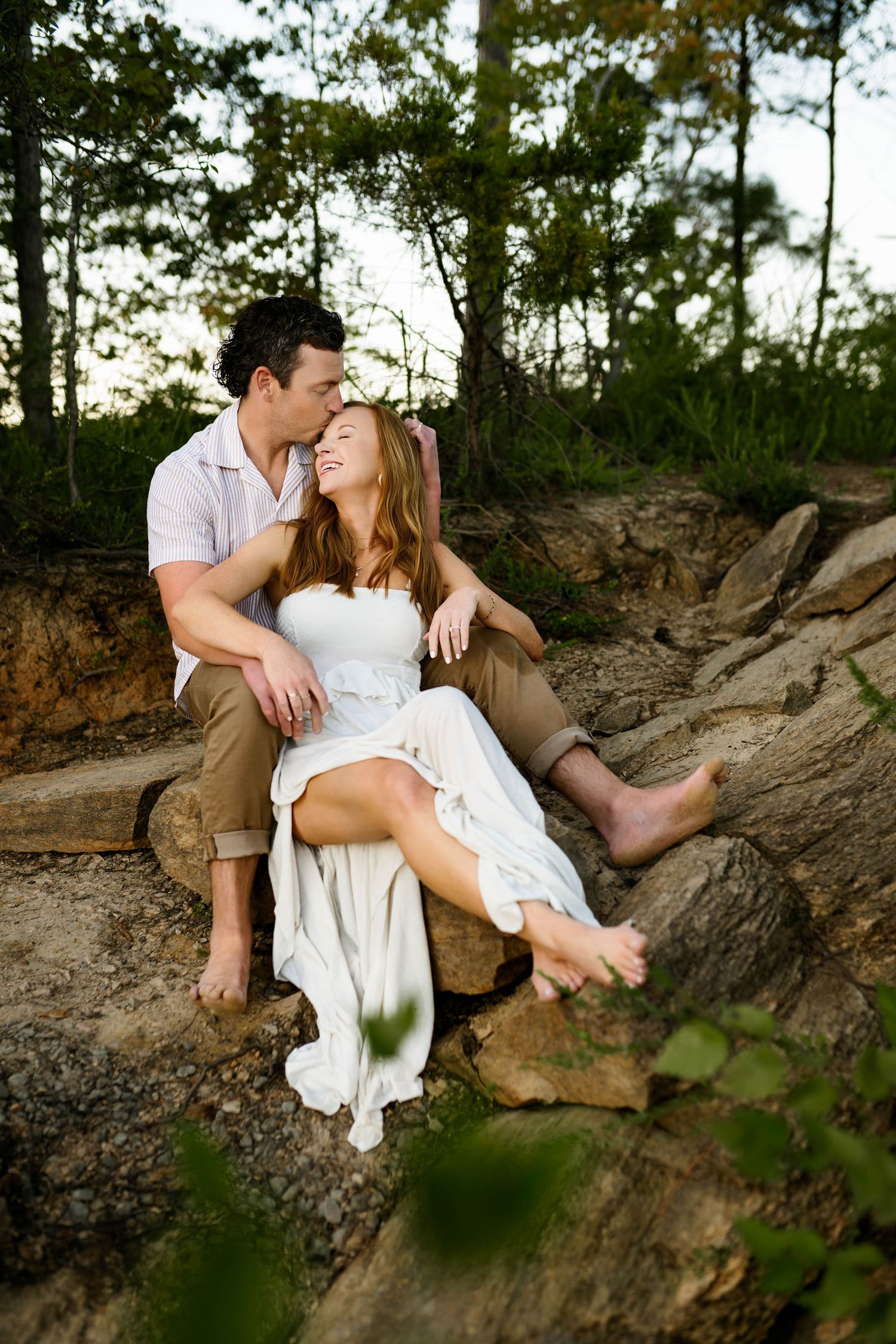 A romantic couple sits together on a rocky outcrop surrounded by trees during an outdoor photo session with Emma Branch Photography