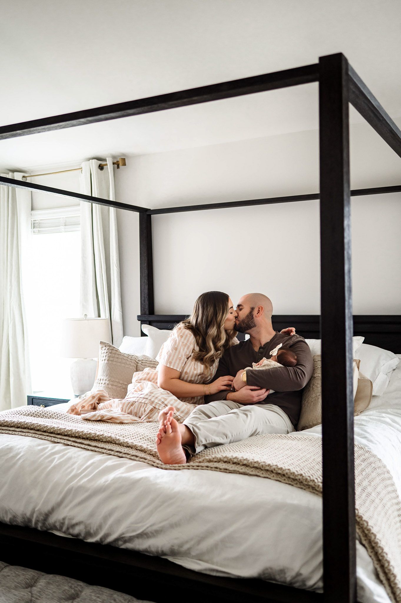 Parents sharing a kiss while sitting on a bed with their newborn baby during a cozy lifestyle newborn photography session