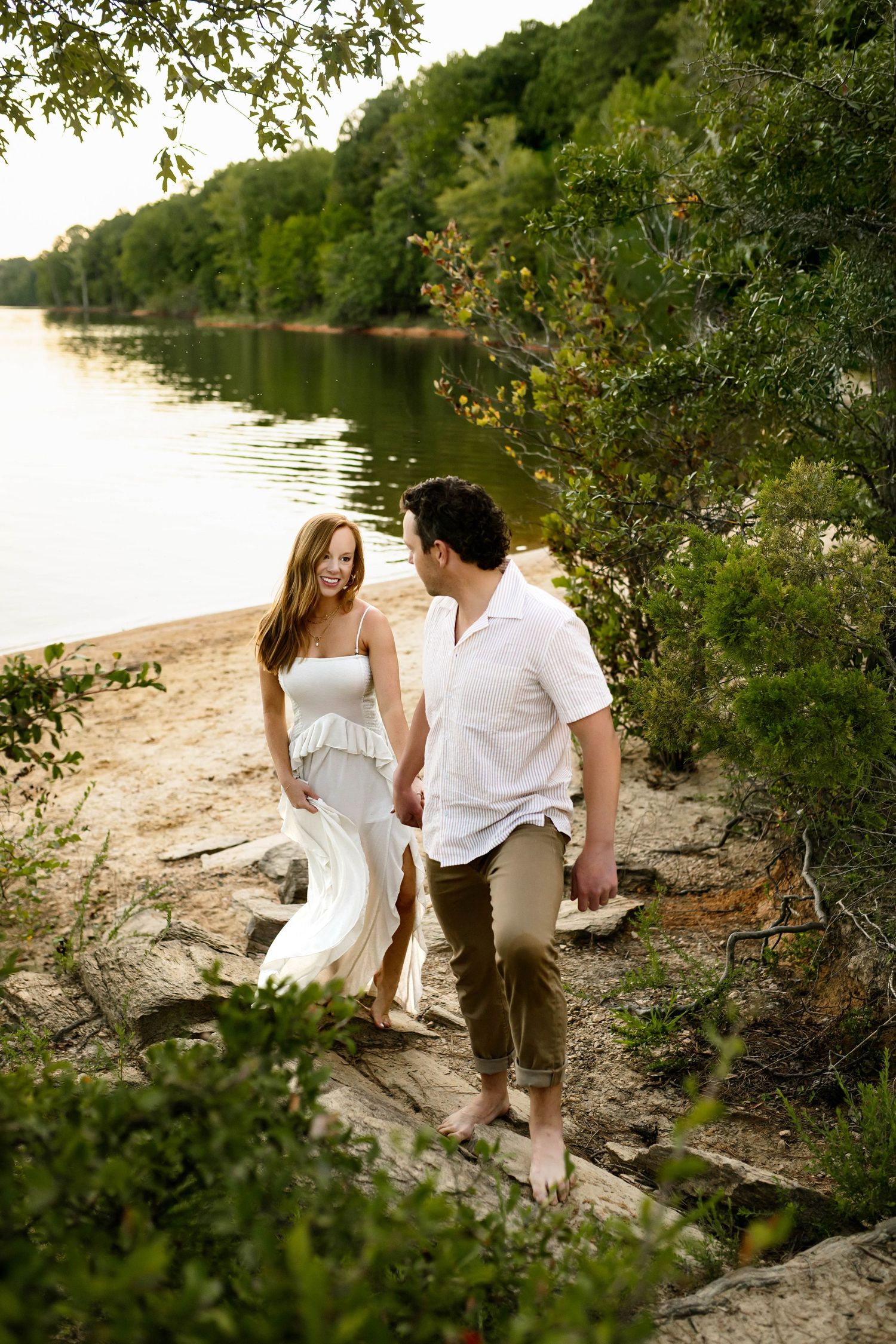 Two people in white clothing share an intimate moment on a sandy riverbank surrounded by lush green foliage with Raleigh family photographer