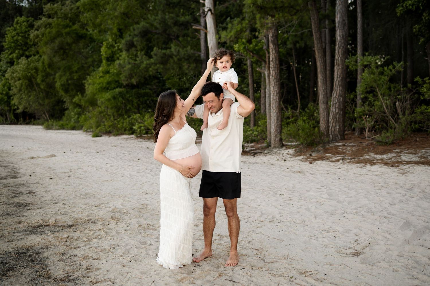 Pregnant mother reaches up playfully towards her young son who sits on father's shoulders during a maternity photography session in Apex NC