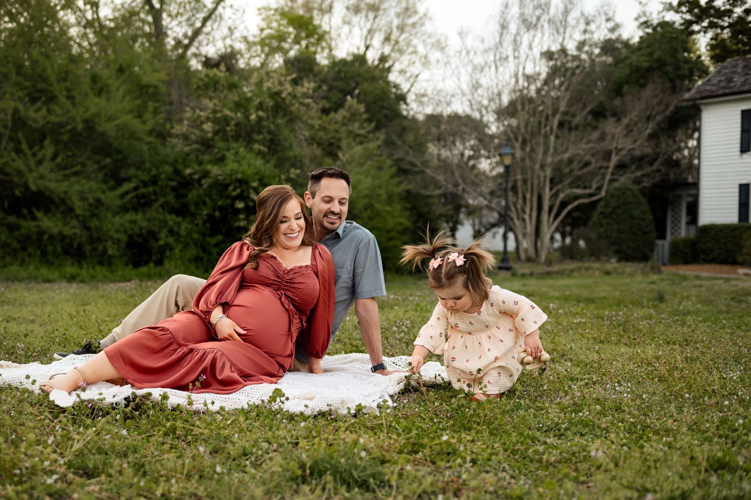 Family sitting on blanket during outdoor Raleigh family photography session while toddler explores nearby