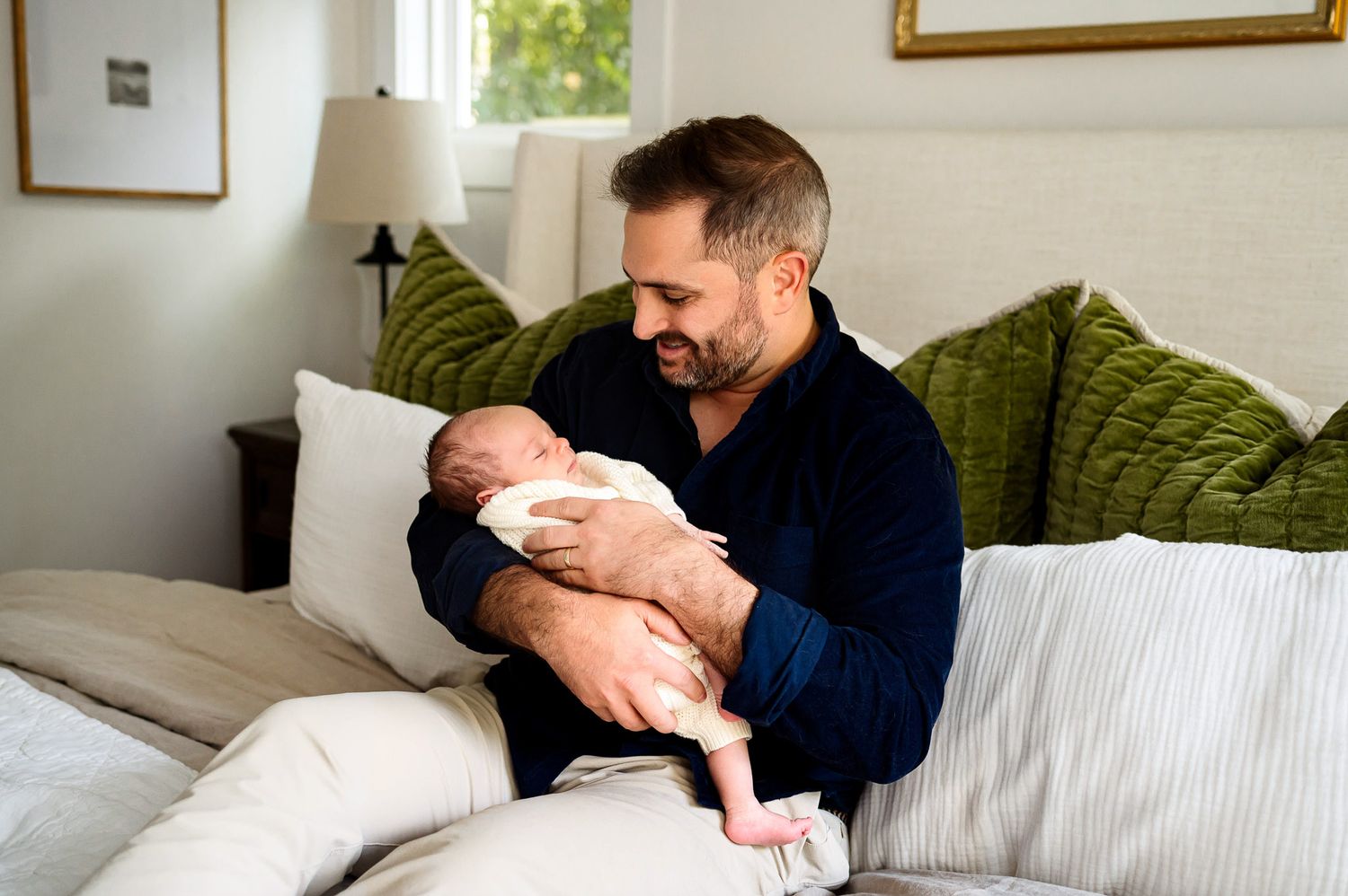 Father holding his newborn baby while sitting on a bed during a relaxed in-home newborn photography session
