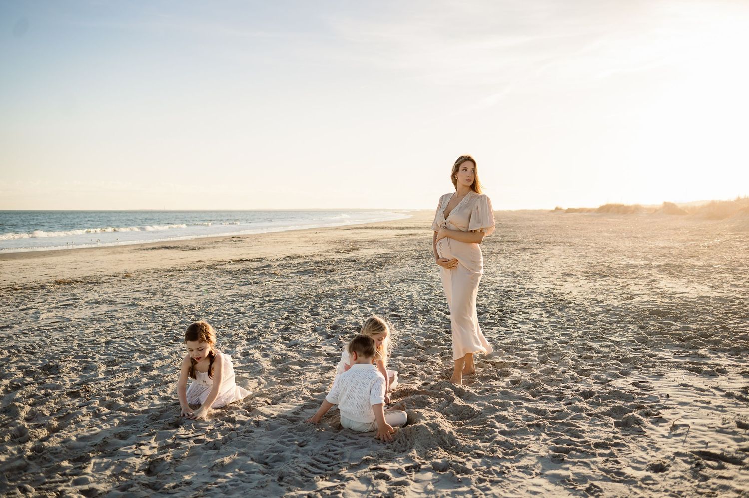Mother holding her belly stands by her young children playing in the sand in a photograph taken by a Raleigh maternity photographer