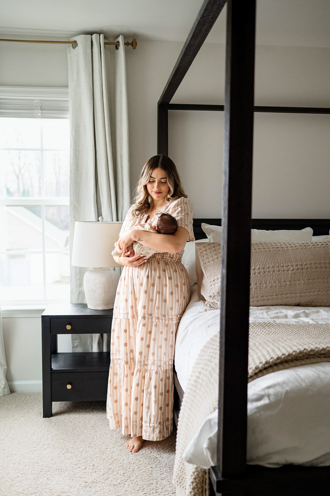 Mother standing beside a window holding her newborn baby during an in-home newborn photography session