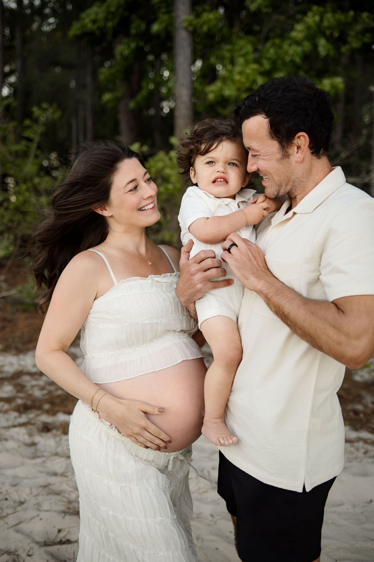 Parents smile at their young son being held by his father while mother holds her exposed pregnant belly during a photography session in Apex