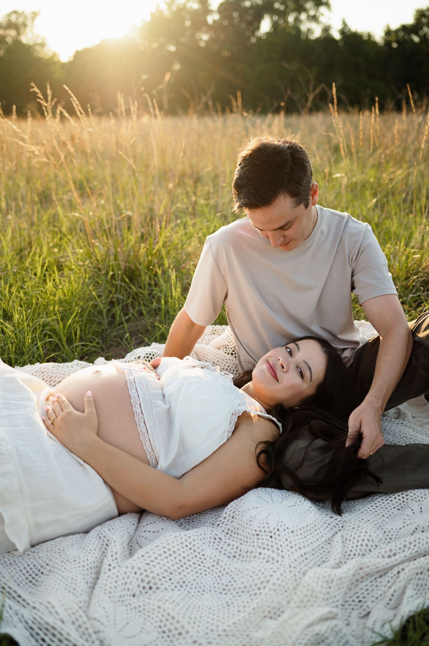 Expecting parents sit together on a blanket in a grassy sunlit field during golden hour with a Raleigh maternity photographer