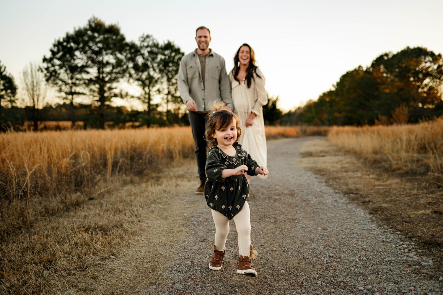 A happy family walks together on a path through golden grasslands at sunset for a candid photo of toddler with raleigh family photographer