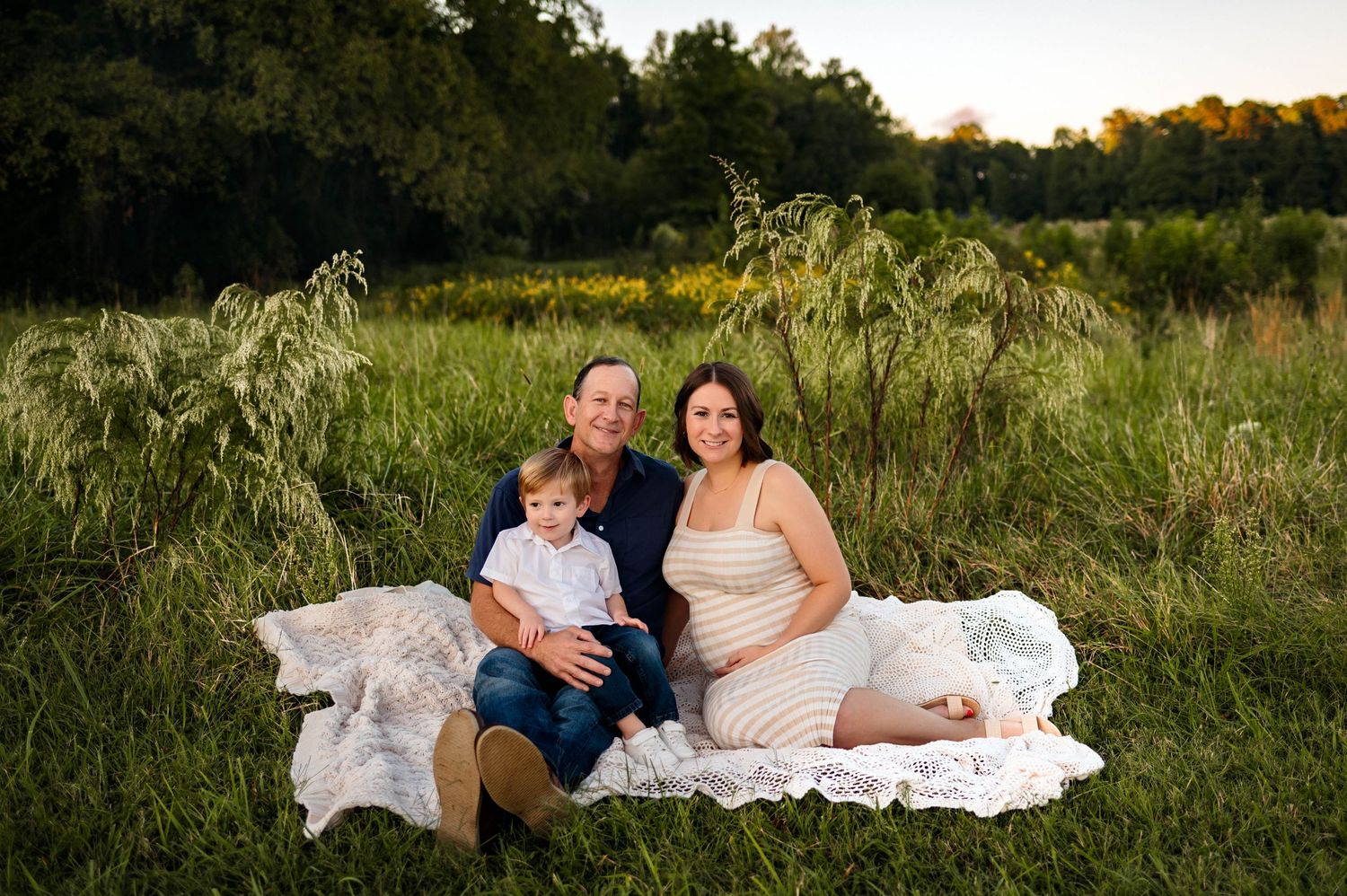 A group sits together on a white blanket in a lush summer field at sunset, surrounded by tall grasses and trees during maternity photography